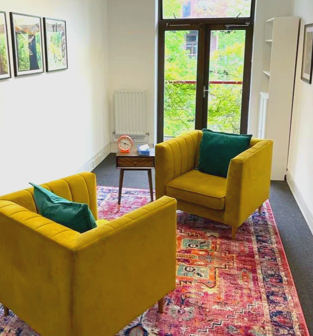 Living room with two yellow armchairs with teal pillows, a small wooden side table with a clock, a colorful area rug, and a large window showing green trees outside.