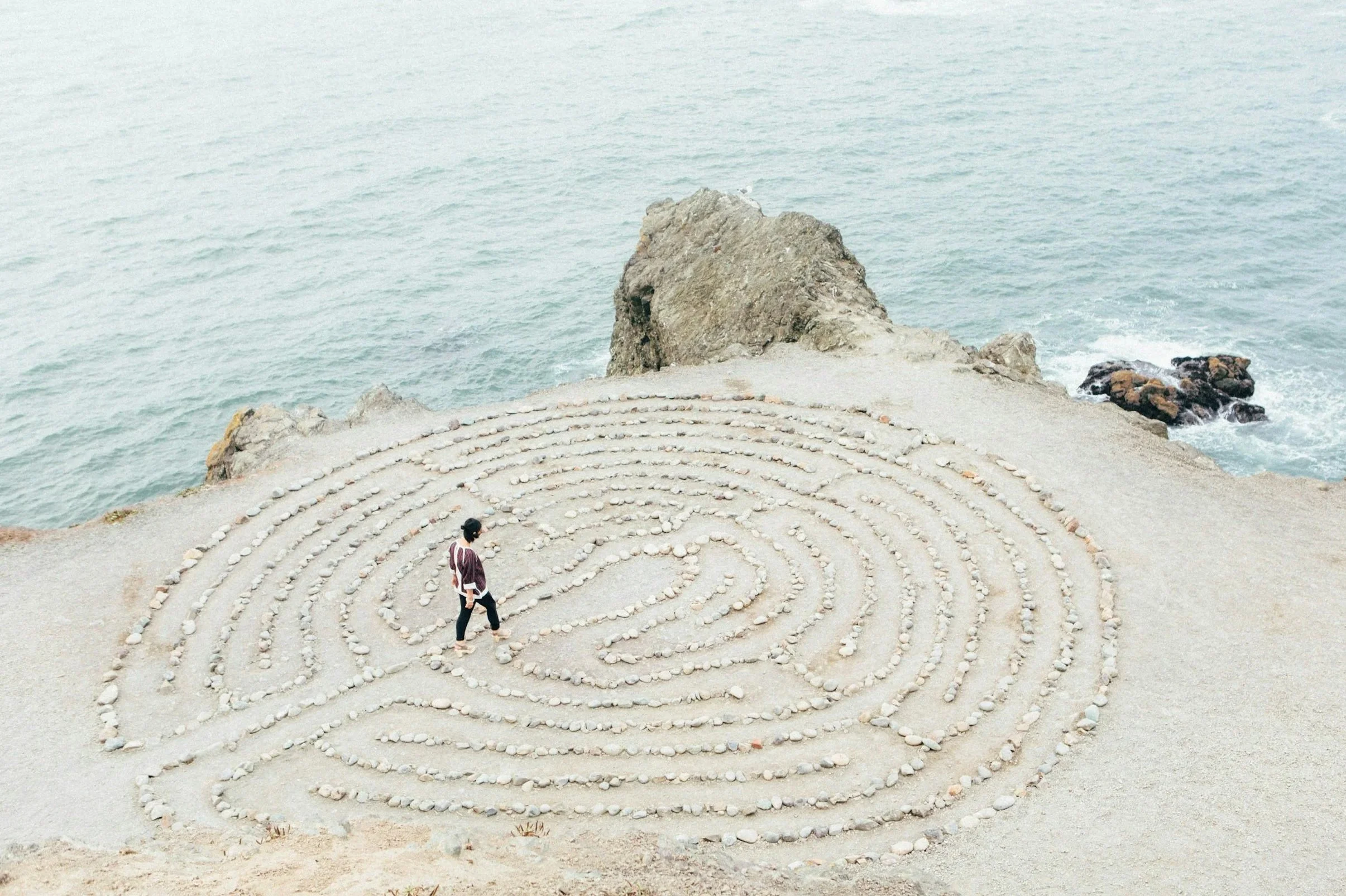 A woman walking through a spiral-shaped stone labyrinth on a sandy beach near the ocean with large rocks in the background.