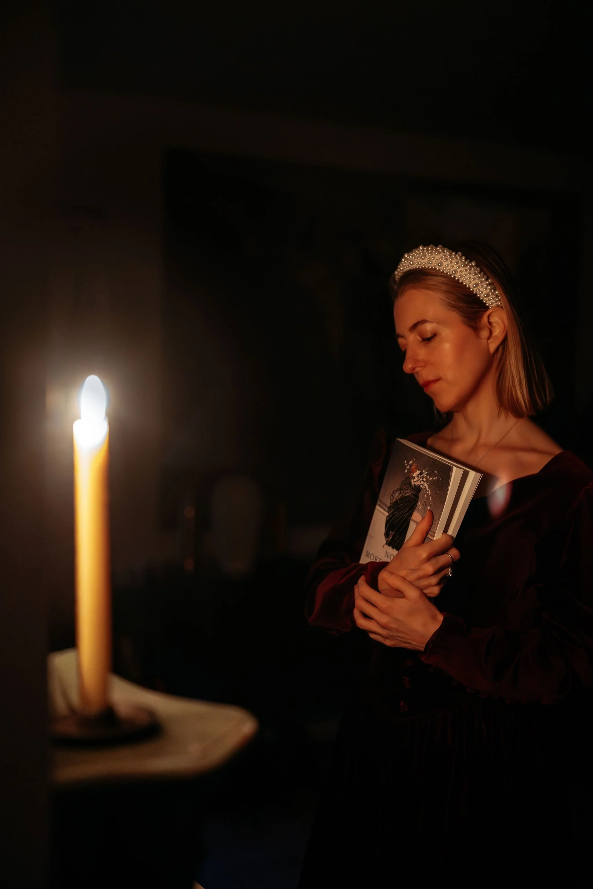 A woman wearing a pearl headband, holding a book, stands near a lit candle in a dark room.
