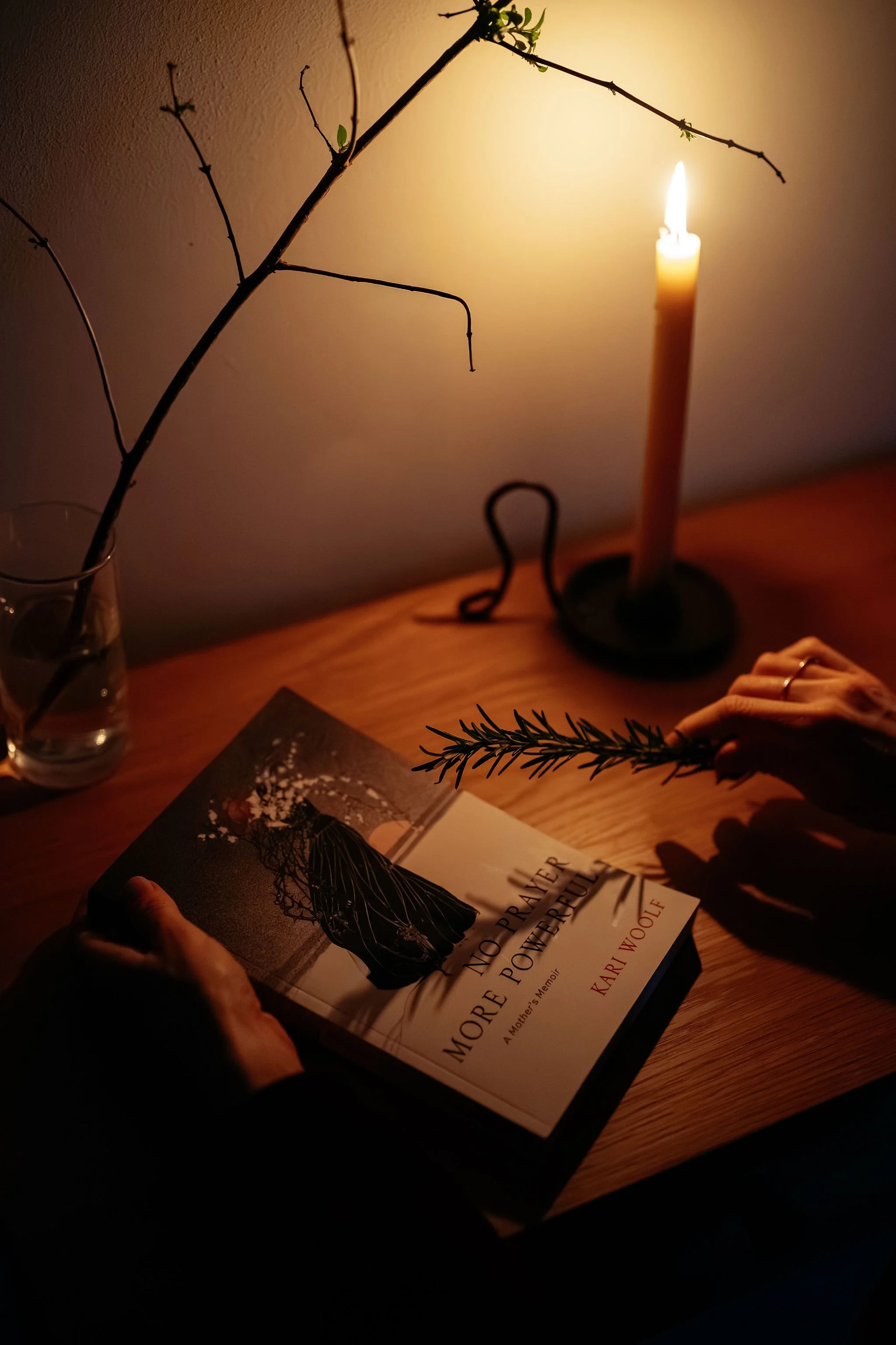 A person holding a sprig of rosemary over a book titled 'More No Prayer More Powerful' in a dimly lit room with a lit candle and a glass with a branch in it on a wooden table.