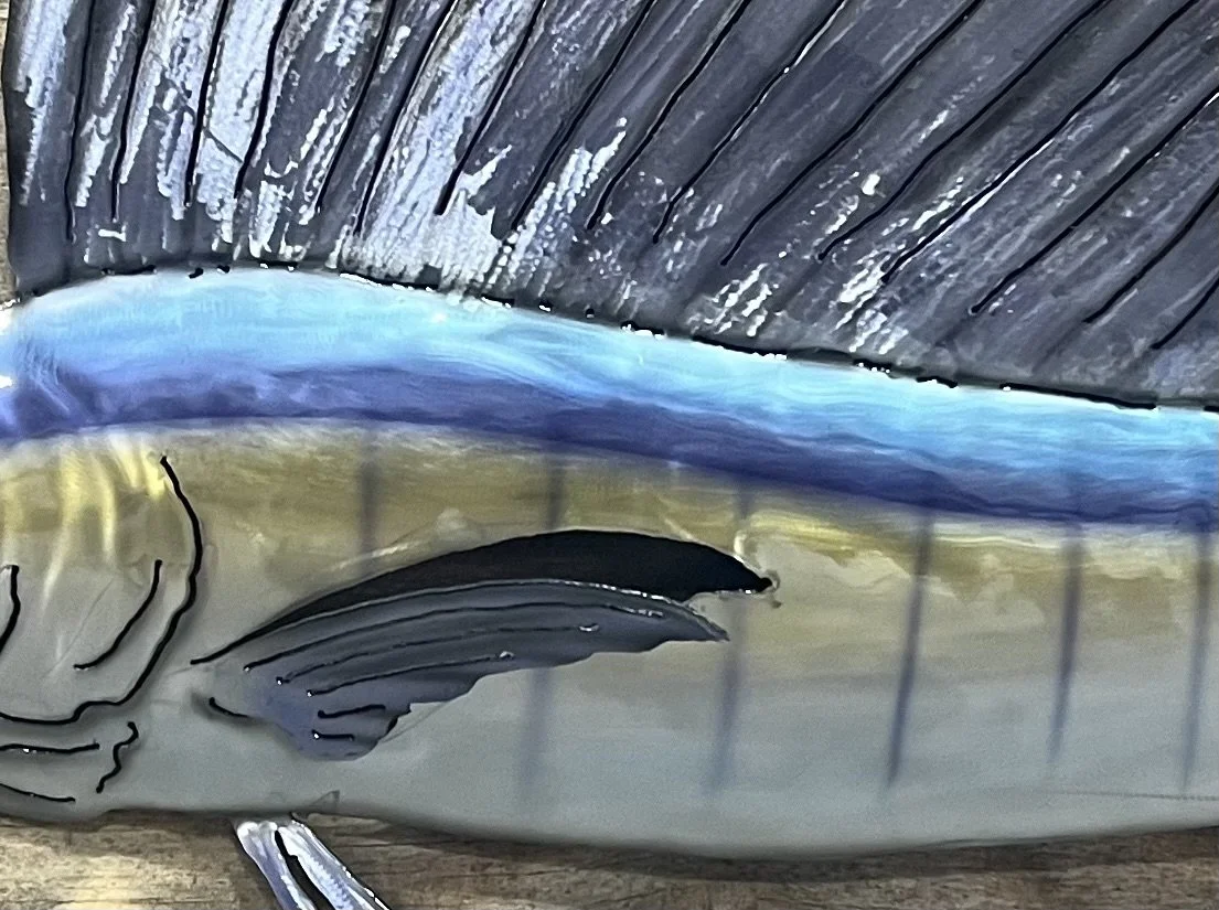 Close-up of a painted fish sculpture, showing detailed scales, fins, and a blue stripe, with a weathered wooden surface beneath.
