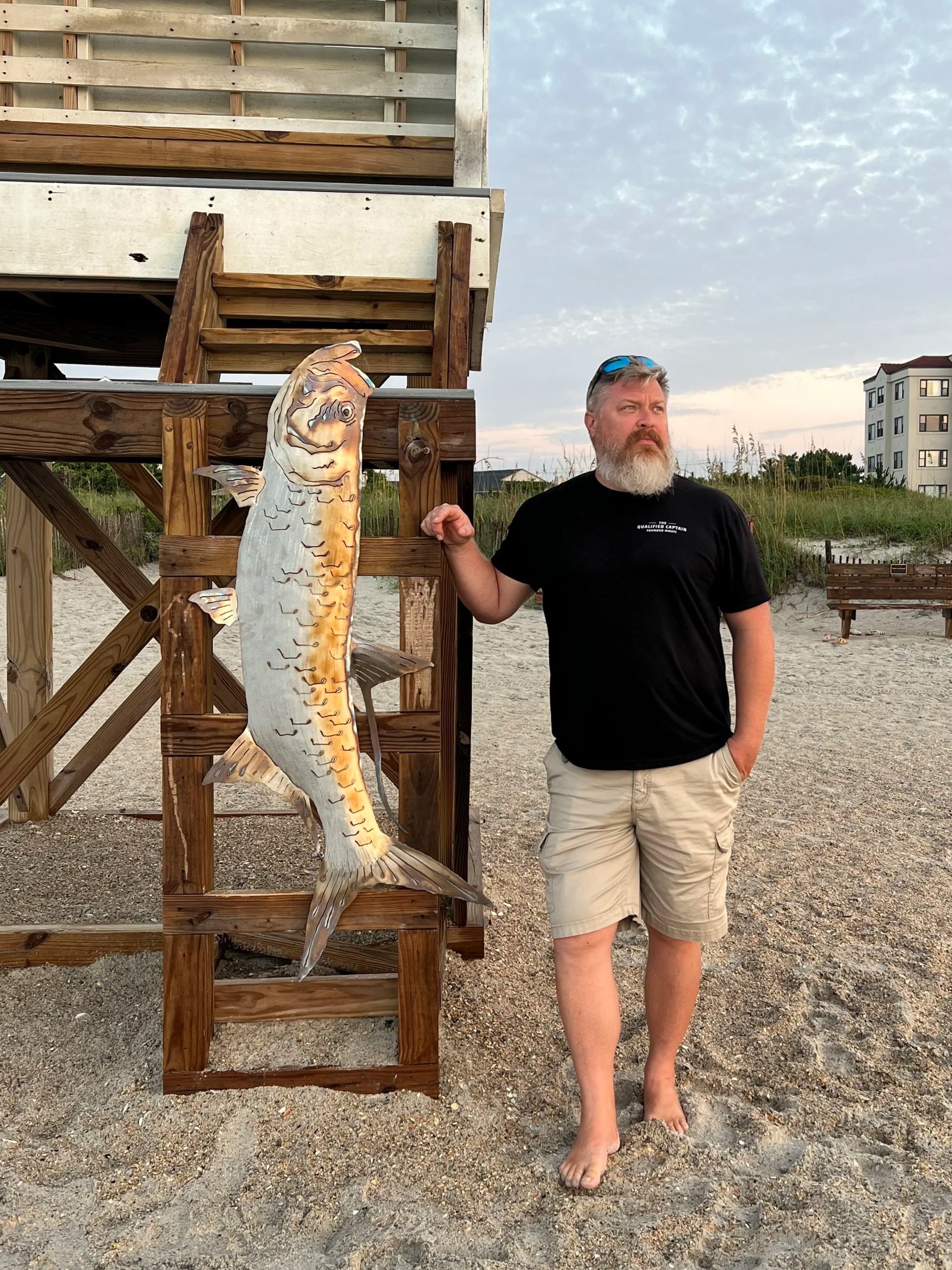 A man with a beard and sunglasses standing barefoot on a sandy beach next to a wooden structure, holding a large fish cutout.