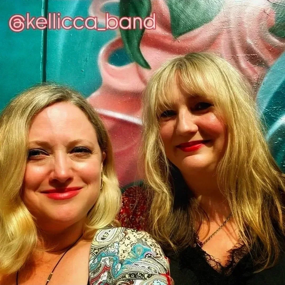 Two blonde women smiling standing Infront of a colourful wall with graffiti art on it