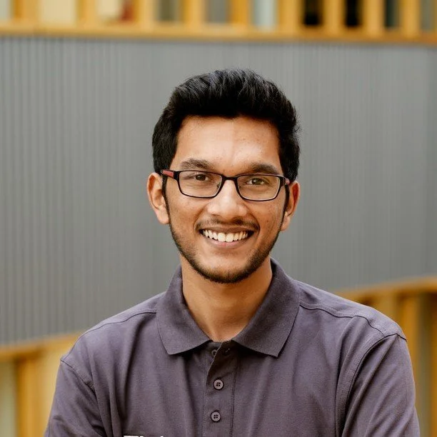A smiling young man with glasses, wearing a dark gray polo shirt, in an indoor setting with bookshelves in the background.