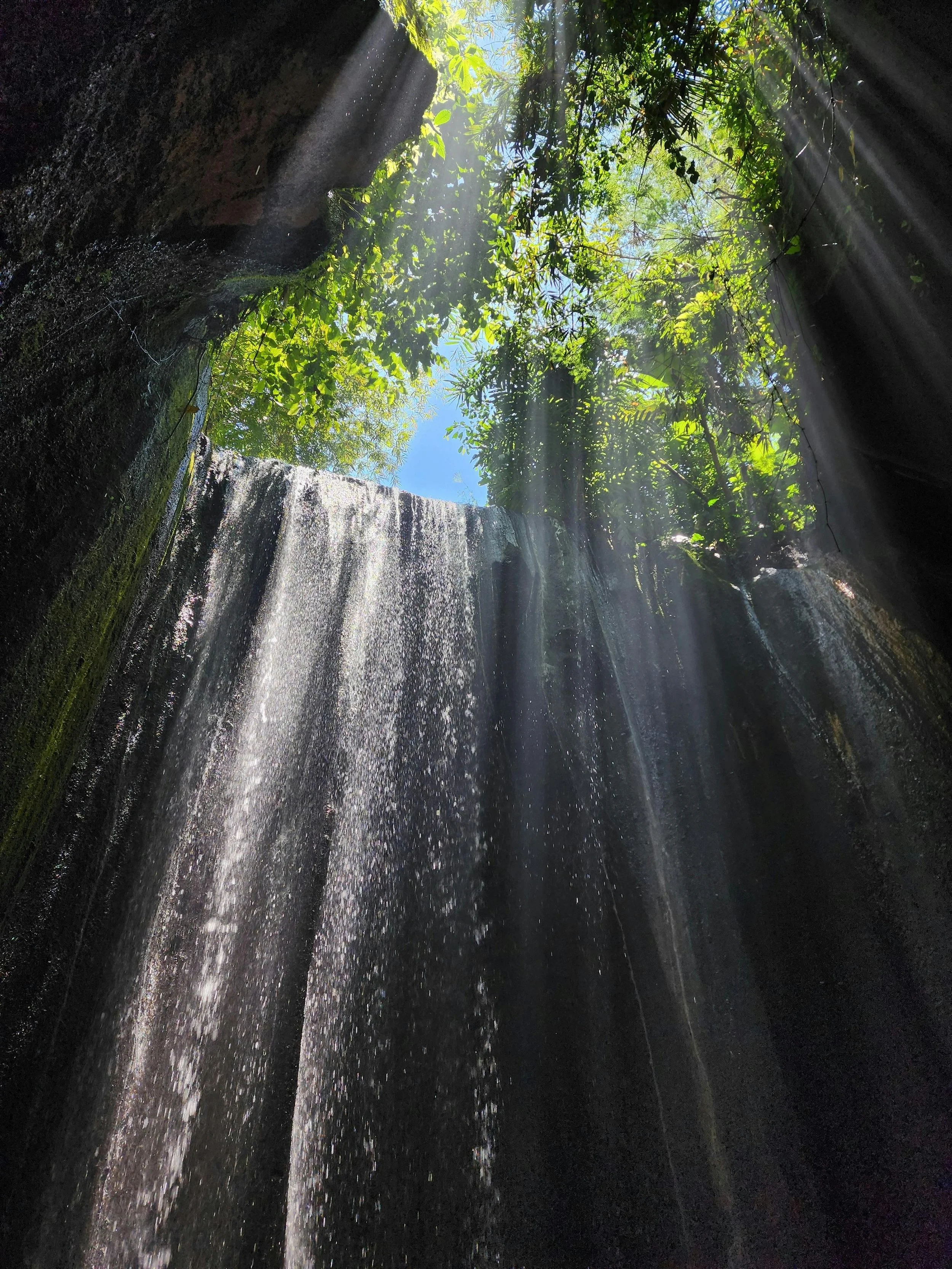 Looking up at a waterfall from the base with sunlight streaming through lush green foliage and blue sky visible above.