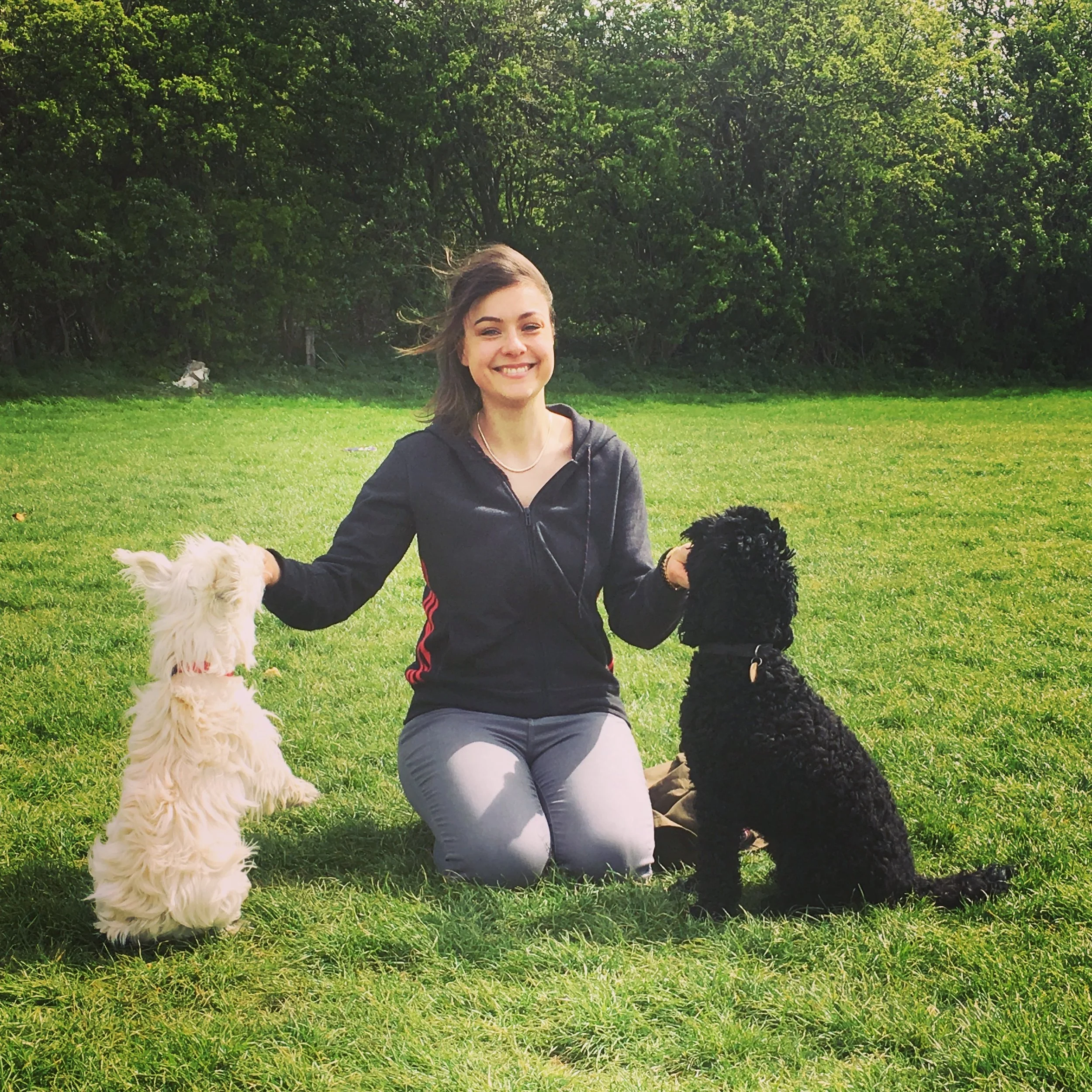 A smiling woman kneels on the grass with two dogs, one white and one black, outdoors with trees in the background.