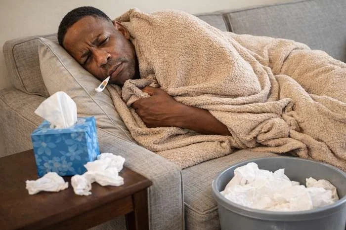 African American man lying on a couch wrapped in a blanket, appearing feverish and ill with tissues and a trash can nearby, illustrating severe kratom withdrawal symptoms.