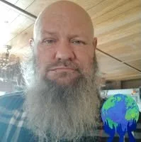 A man with a bald head and a long gray beard looking at the camera, with a small globe in the bottom right corner.