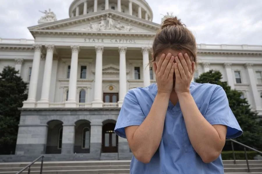 Healthcare worker in medical scrubs standing on the steps of a state capitol building with her face in her hands, expressing concern over a legislative decision affecting public health.