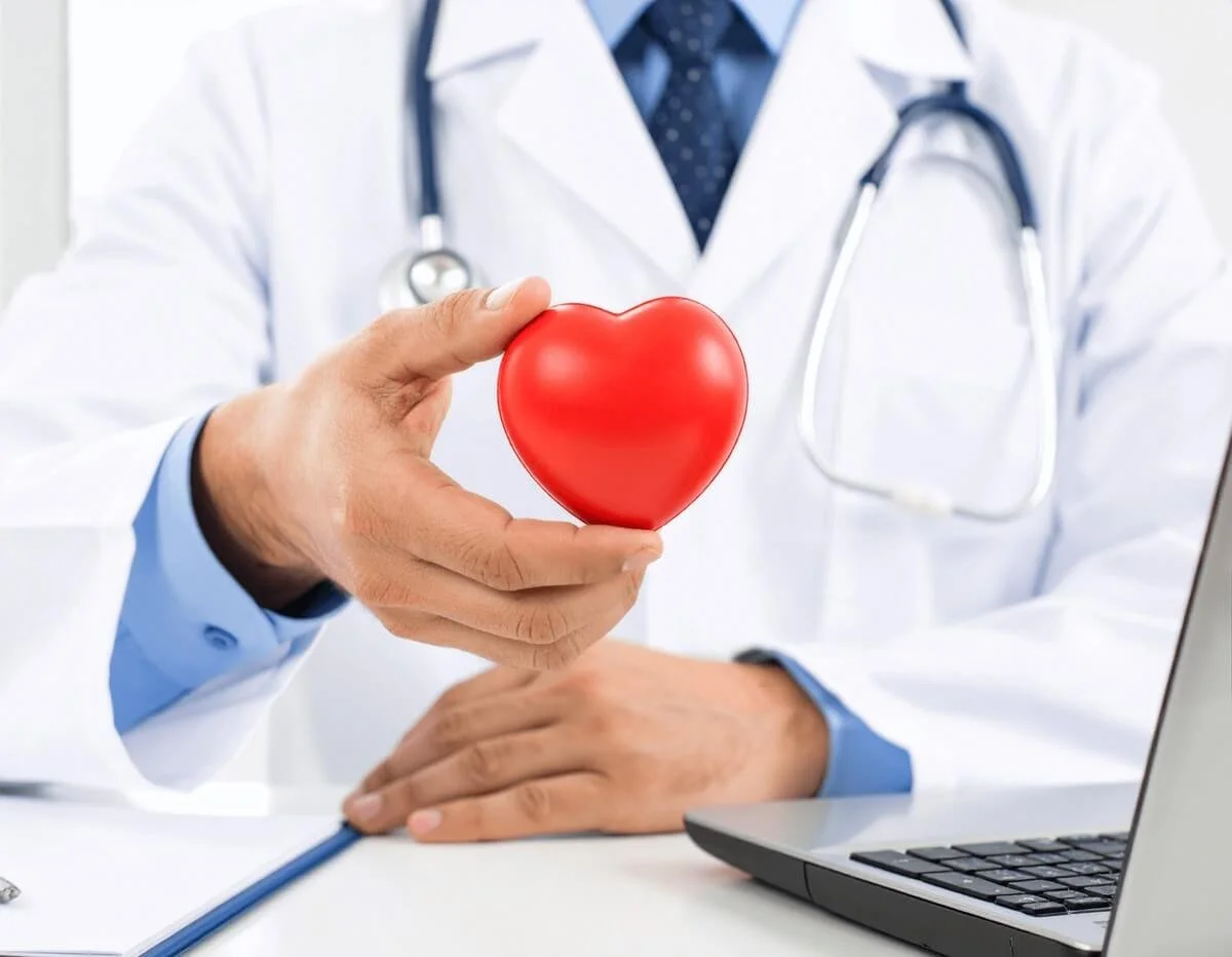 Doctor in white lab coat with stethoscope holding a red heart at a desk next to a laptop