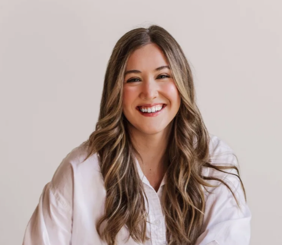 A woman with long, wavy, brown hair smiling at the camera, wearing a white blouse against a plain background.