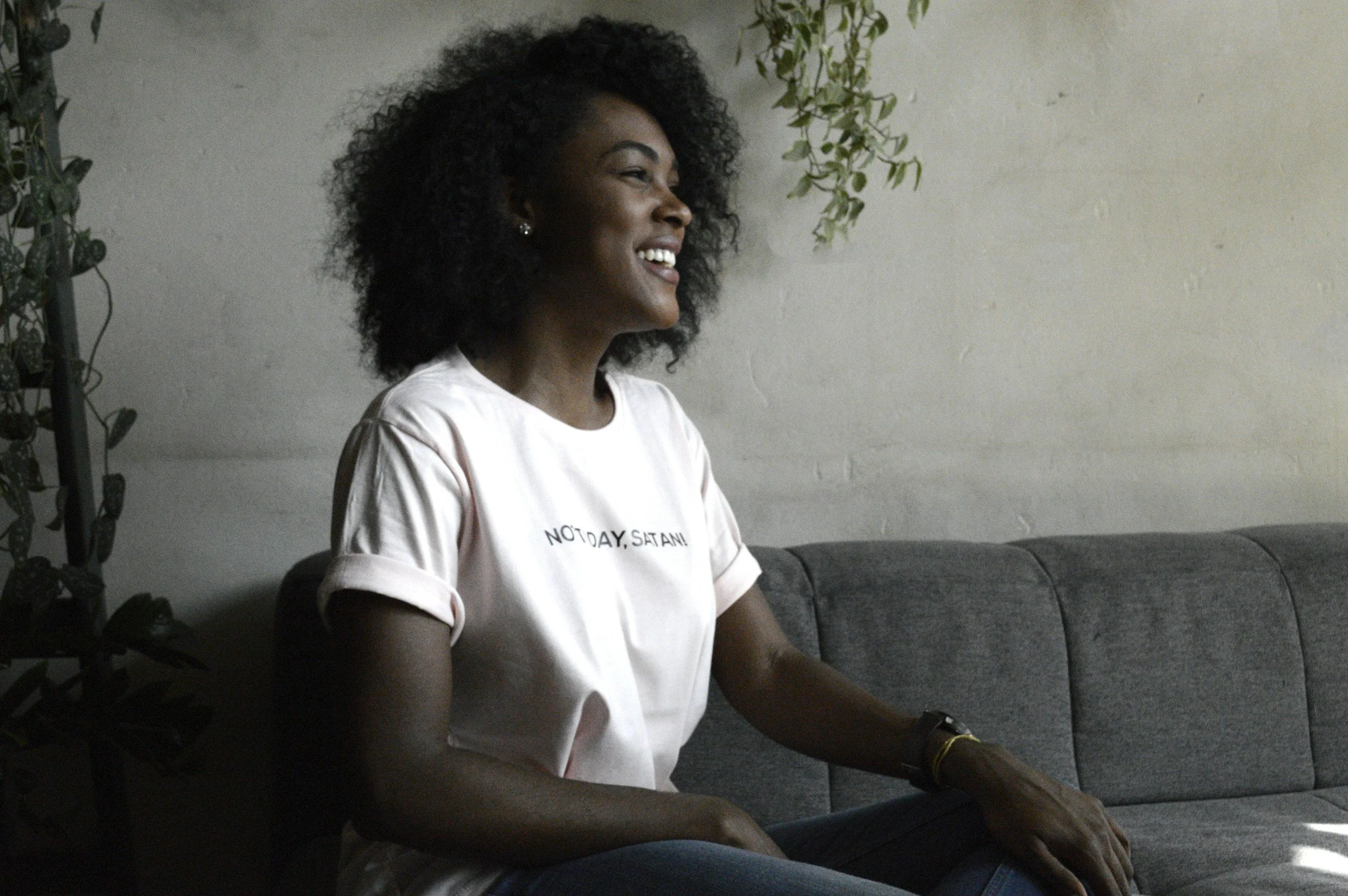 A woman with curly black hair, smiling and sitting on a gray couch in a room with a white brick wall and some greenery.