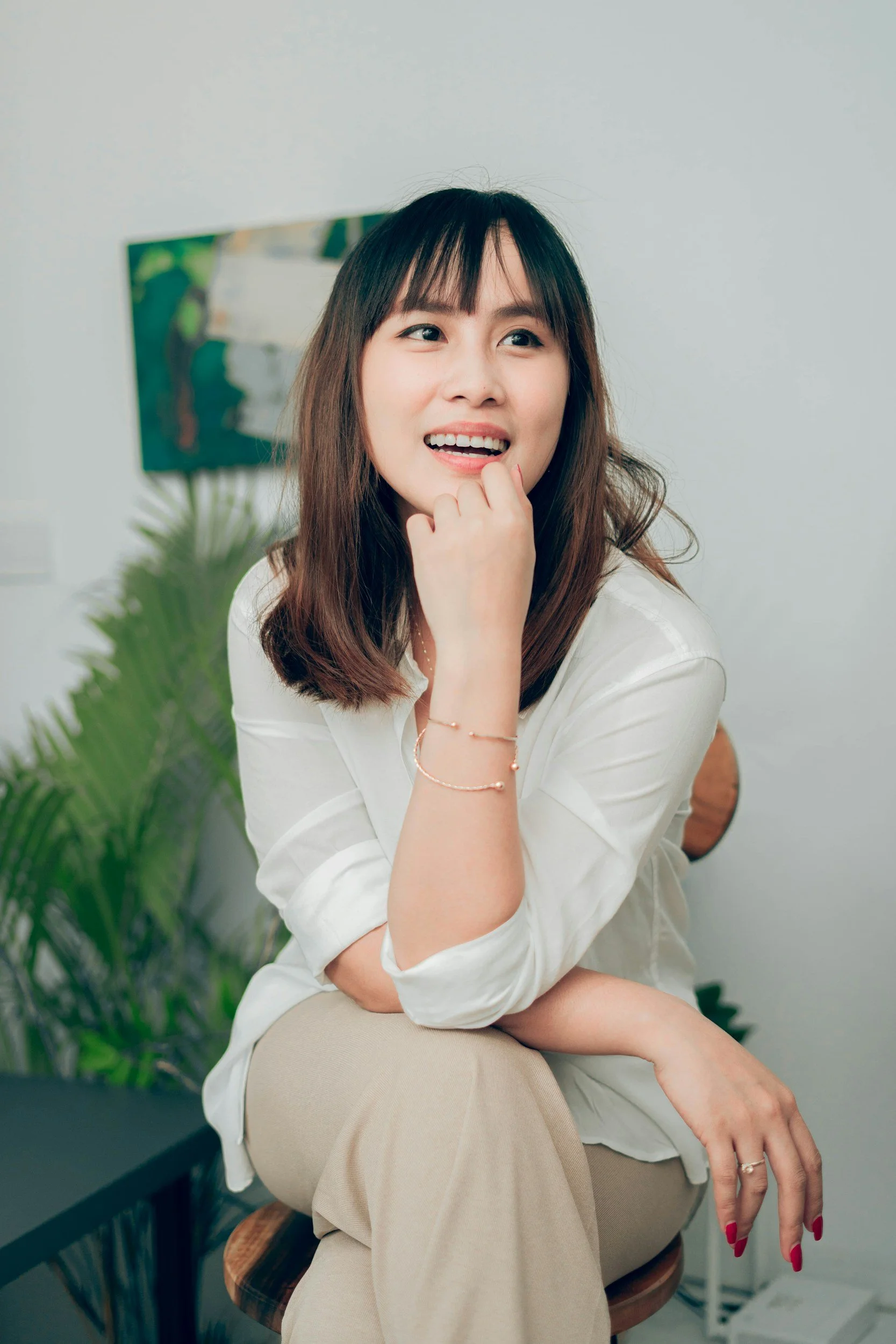 A woman with brown hair and bangs, wearing a white top and beige pants, sitting on a chair with her hand near her chin, smiling and looking to the side.
