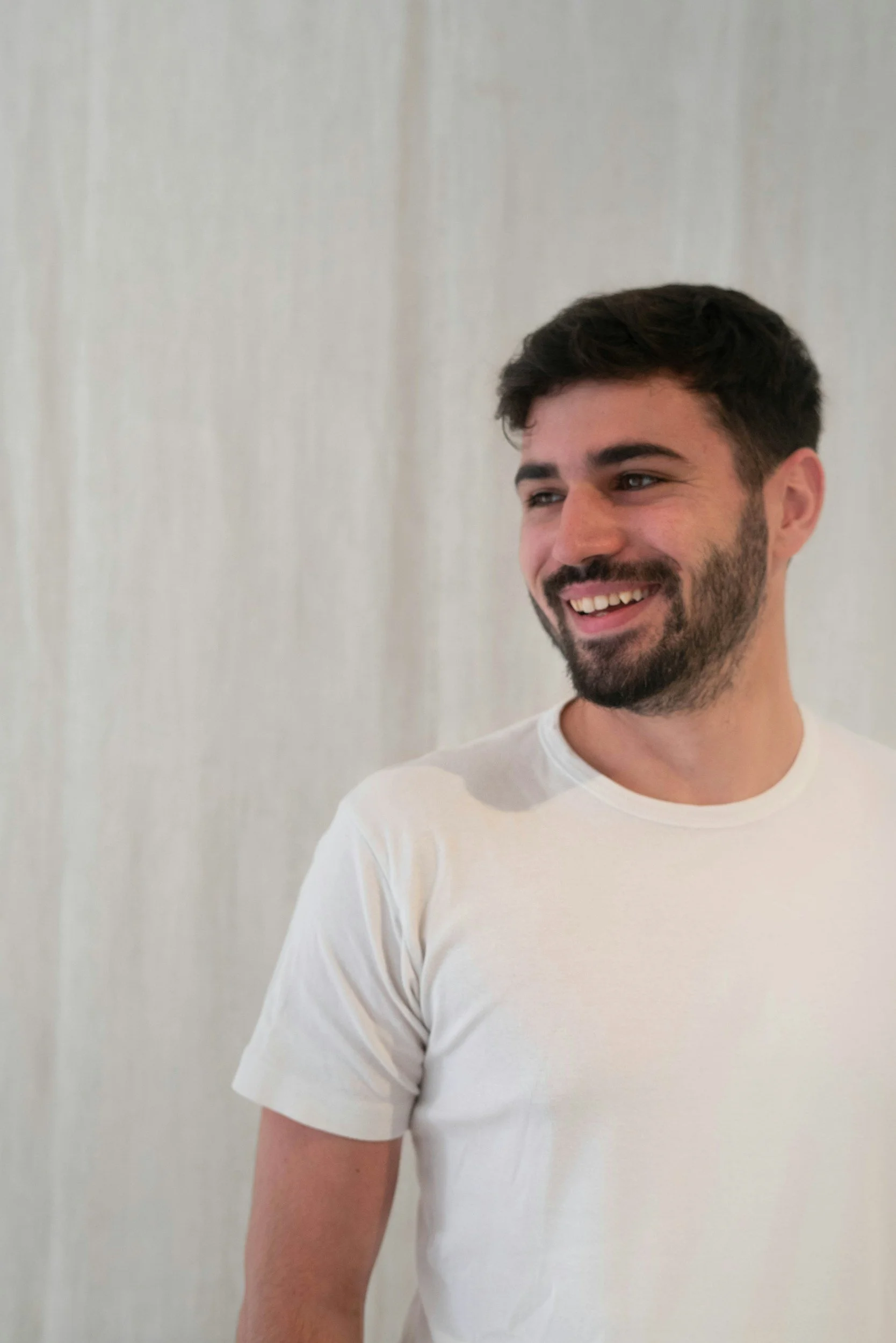A smiling man with dark hair and a beard, wearing a white t-shirt, standing against a light-colored background.
