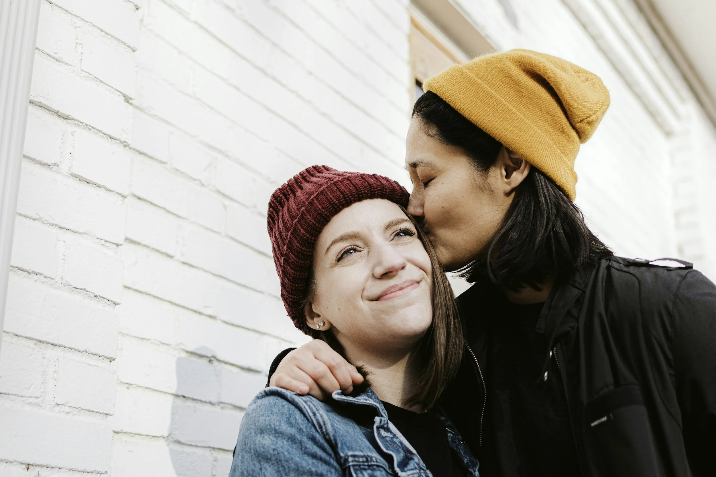 A woman with dark hair wearing an orange beanie kisses a smiling young woman with brown hair and a red beanie on the forehead, against a white brick wall.
