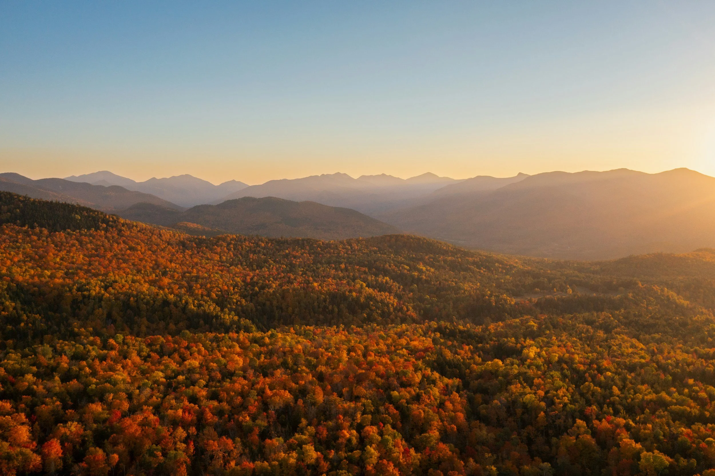 Sunset over a vast mountainous landscape with colorful autumn trees in the foreground.