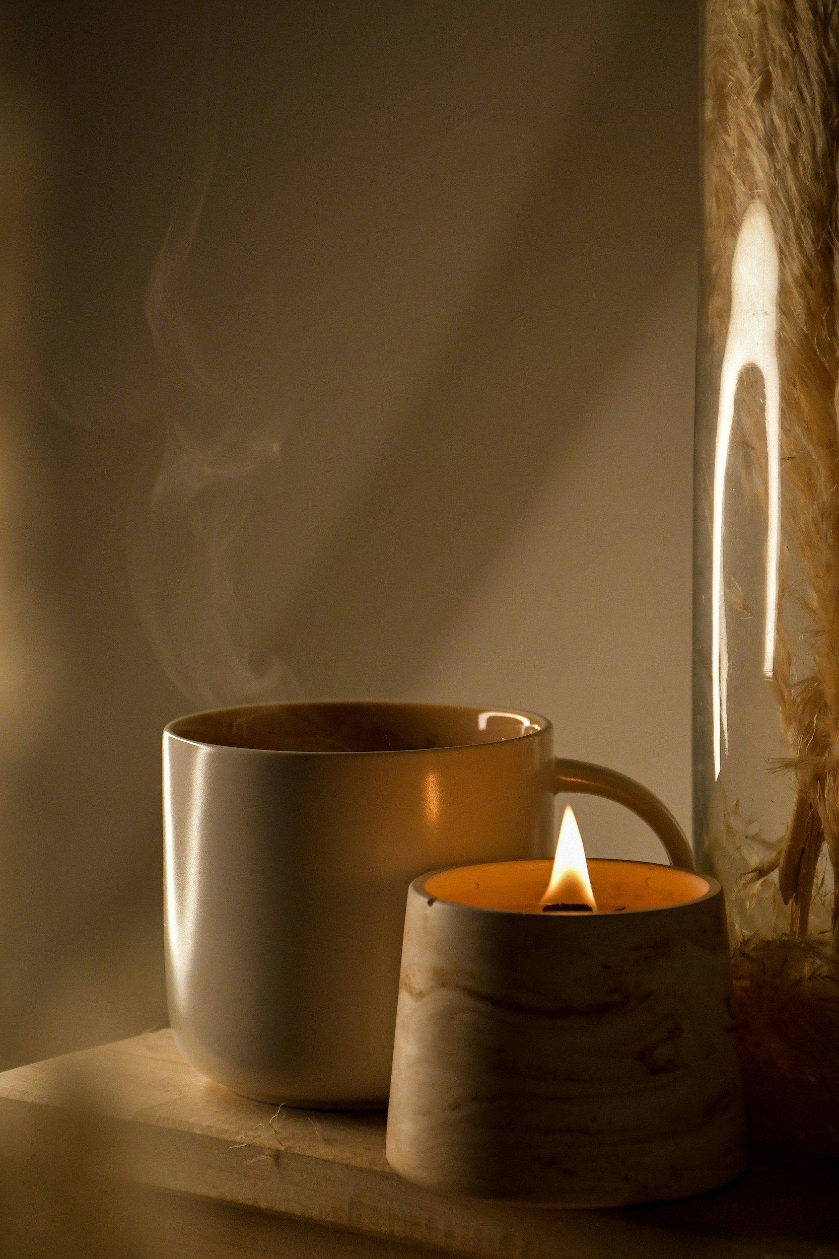 A lit candle and a steaming mug on a wooden surface with a soft, warm glow.