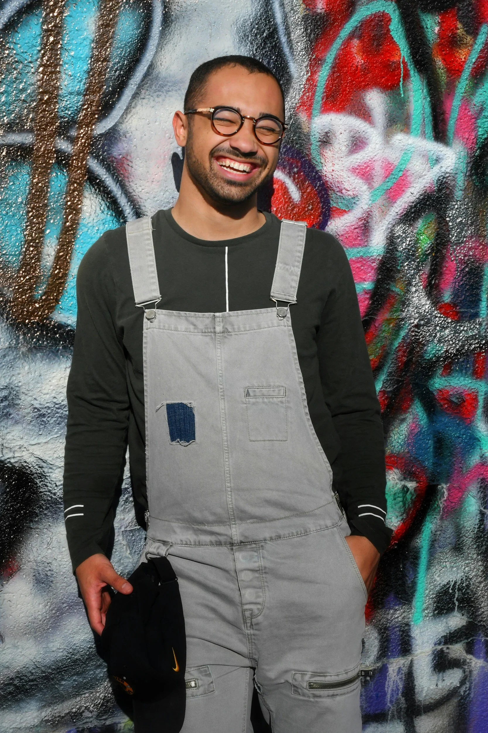 A man with glasses, a beard, and short hair smiling, wearing gray overalls over a black long-sleeve shirt, standing in front of a colorful graffiti wall.