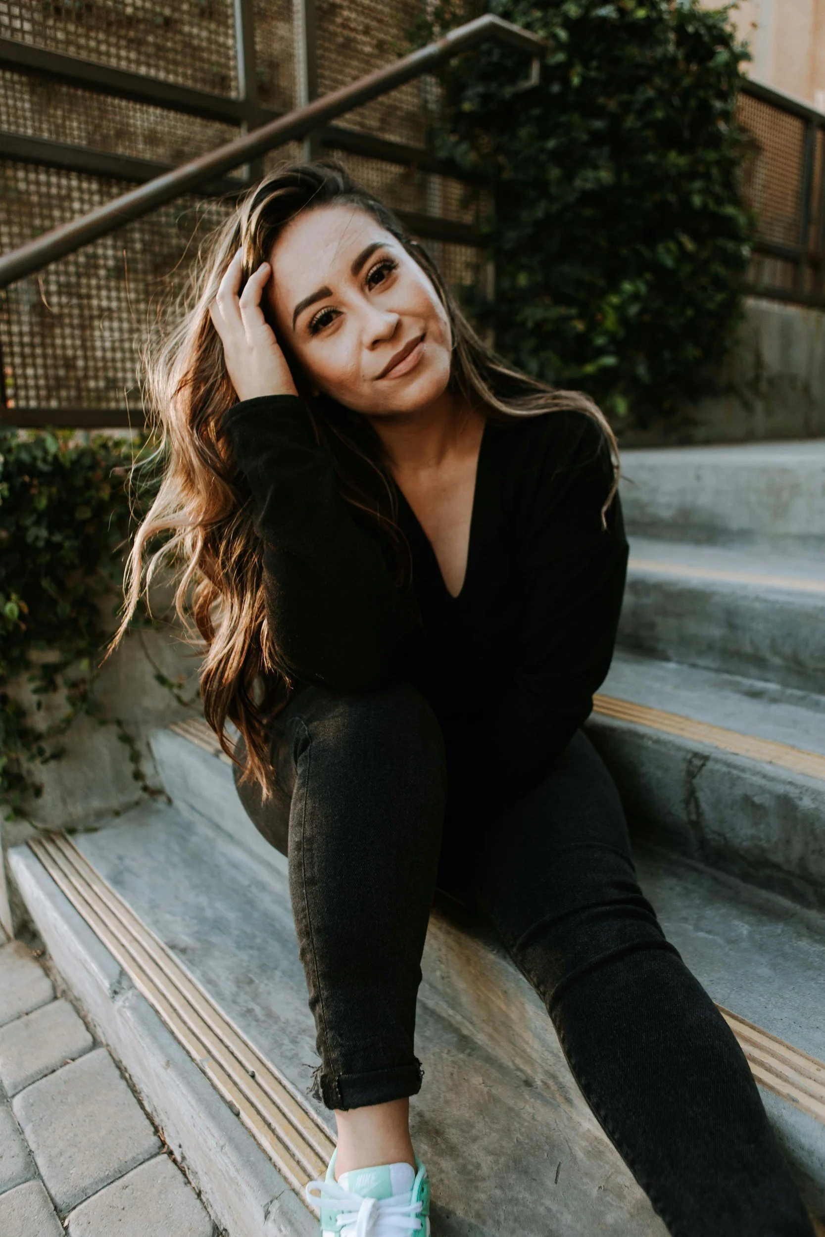 A woman with long wavy brown hair, dressed in a black top and black jeans, sitting on outdoor concrete stairs, smiling softly at the camera, with one hand touching her head, and wearing mint green sneakers.