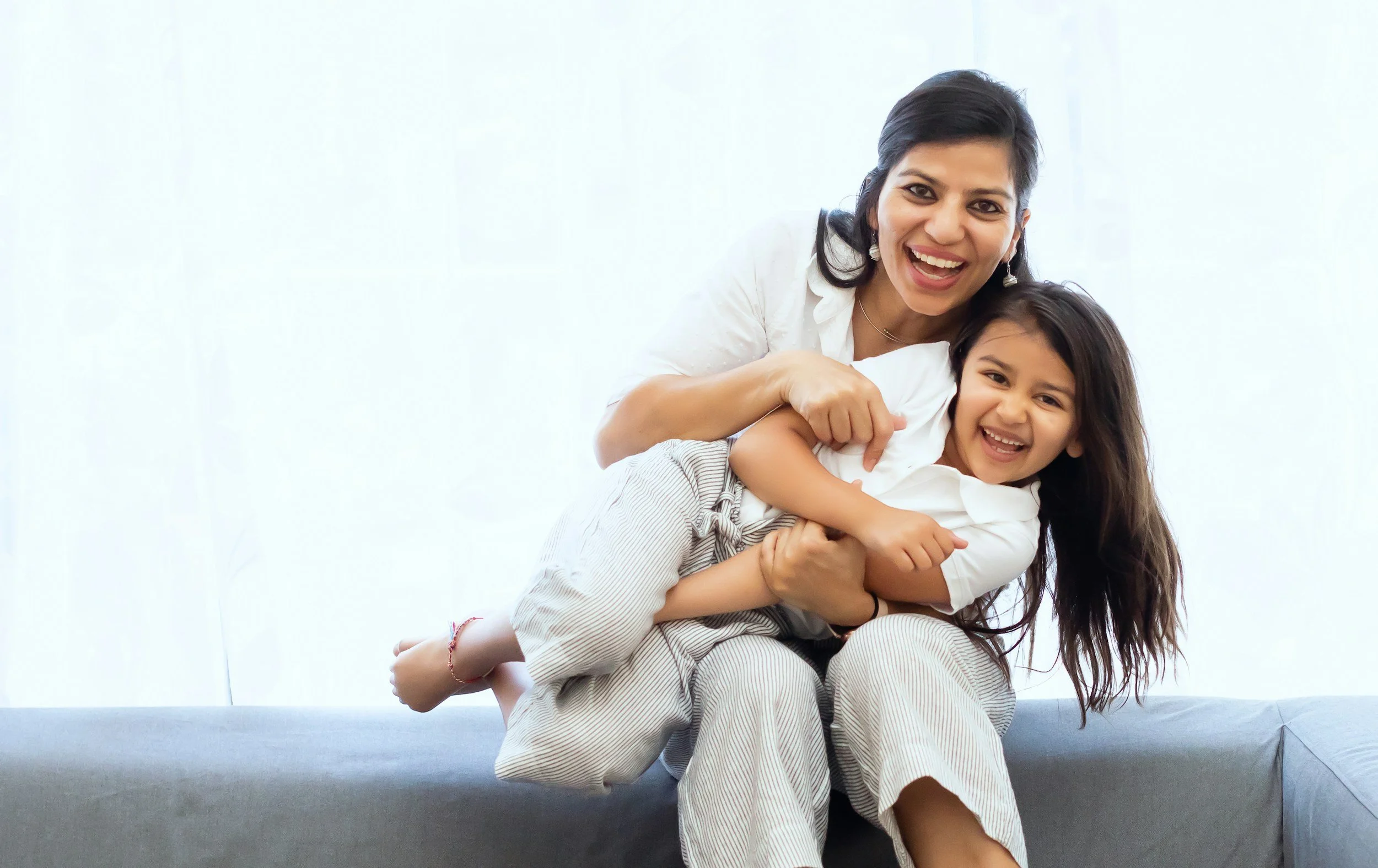 Smiling woman and young girl playing and laughing together on a sofa in a bright room.