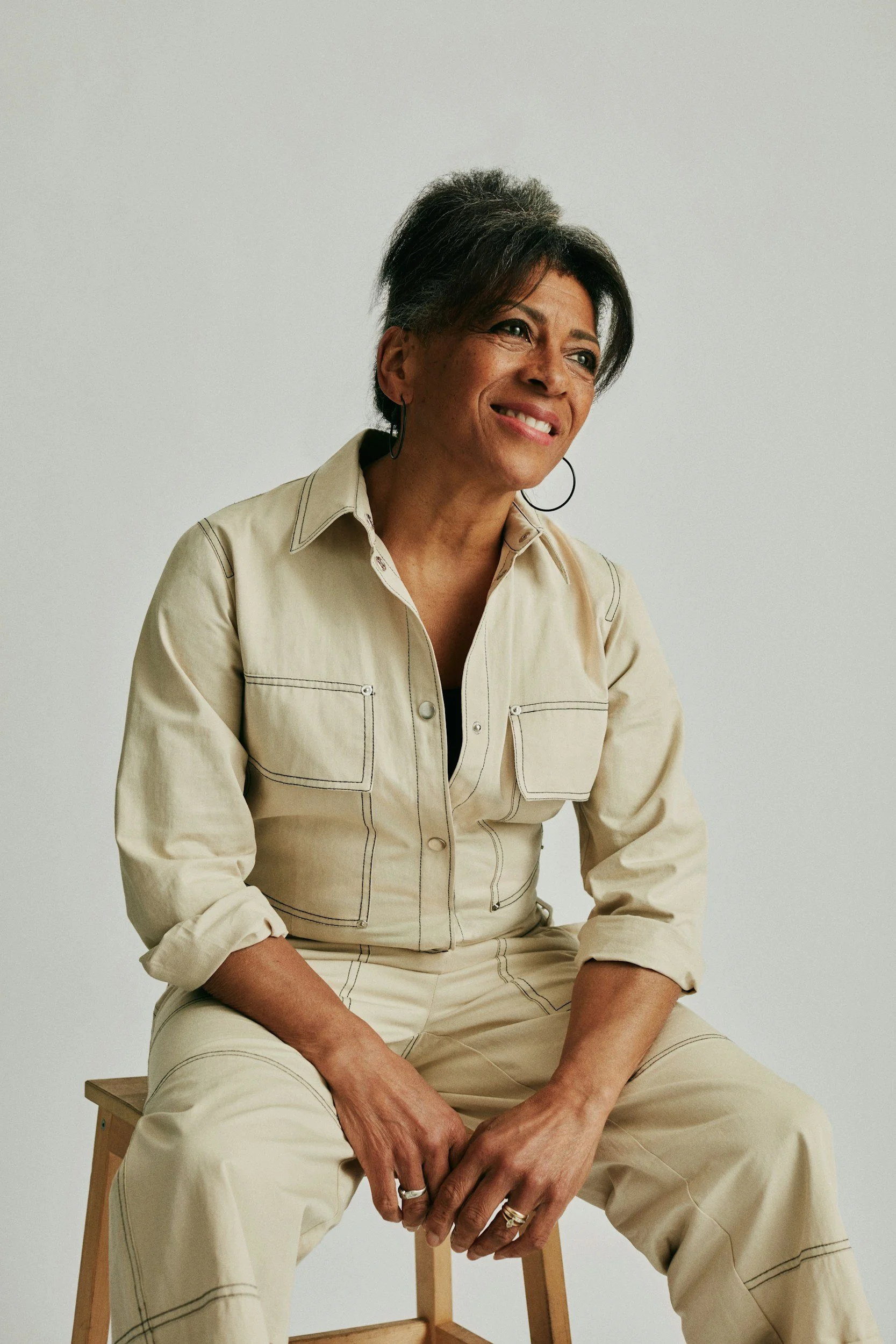 A woman with short, gray hair sitting on a wooden stool against a plain white background. She is wearing a beige utility jacket and matching pants, smiling softly and looking slightly to the side.