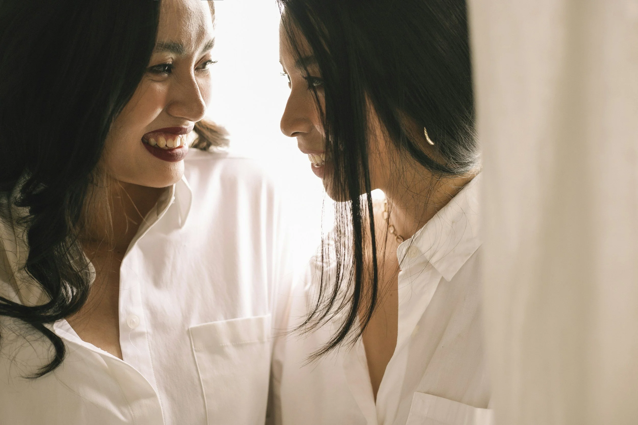 Two women face each other smiling, both wearing white shirts, with dark hair and light skin, in a close-up indoor setting.
