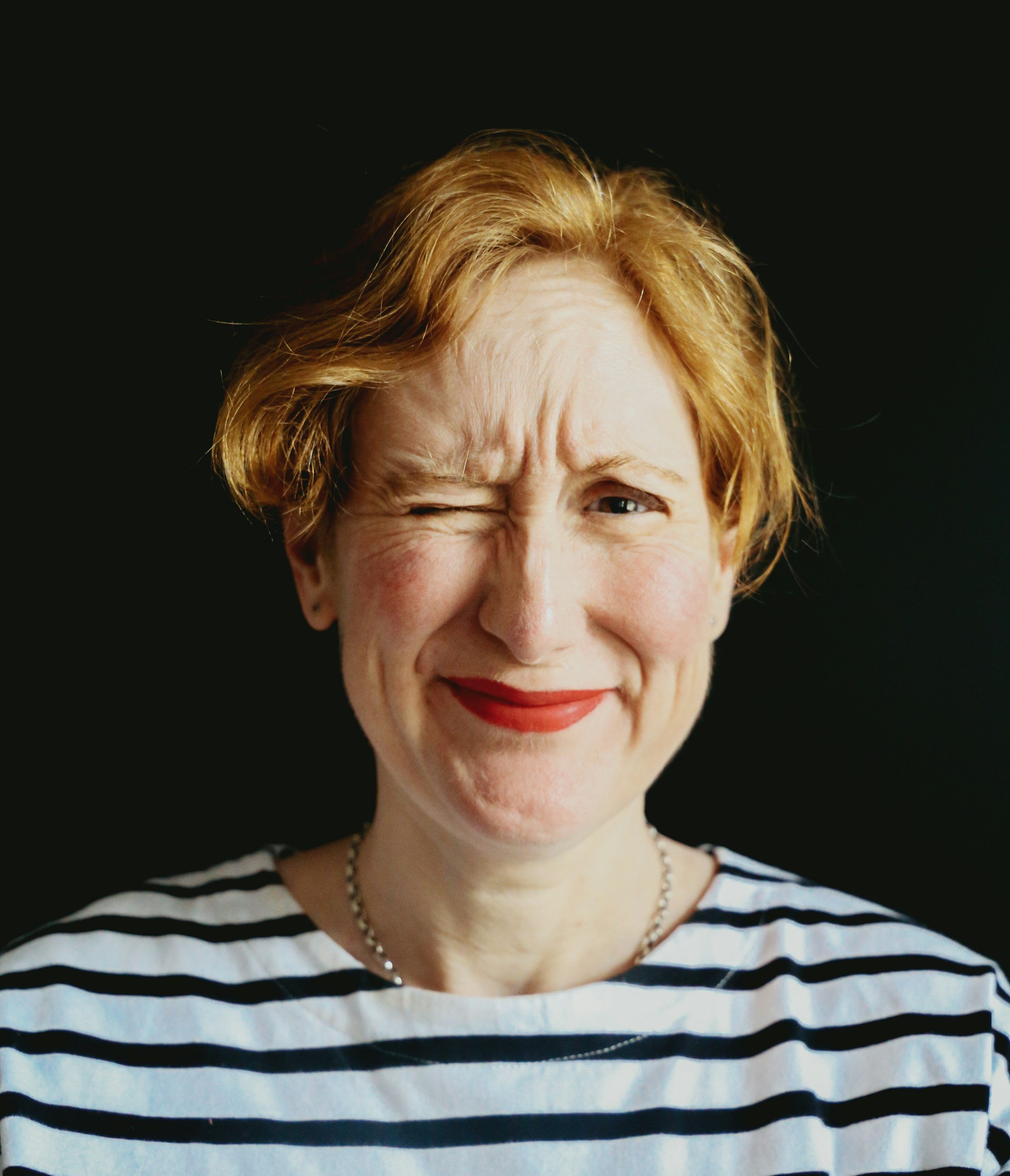 A woman with short red hair winks and smiles, wearing a striped shirt and red lipstick, against a black background.