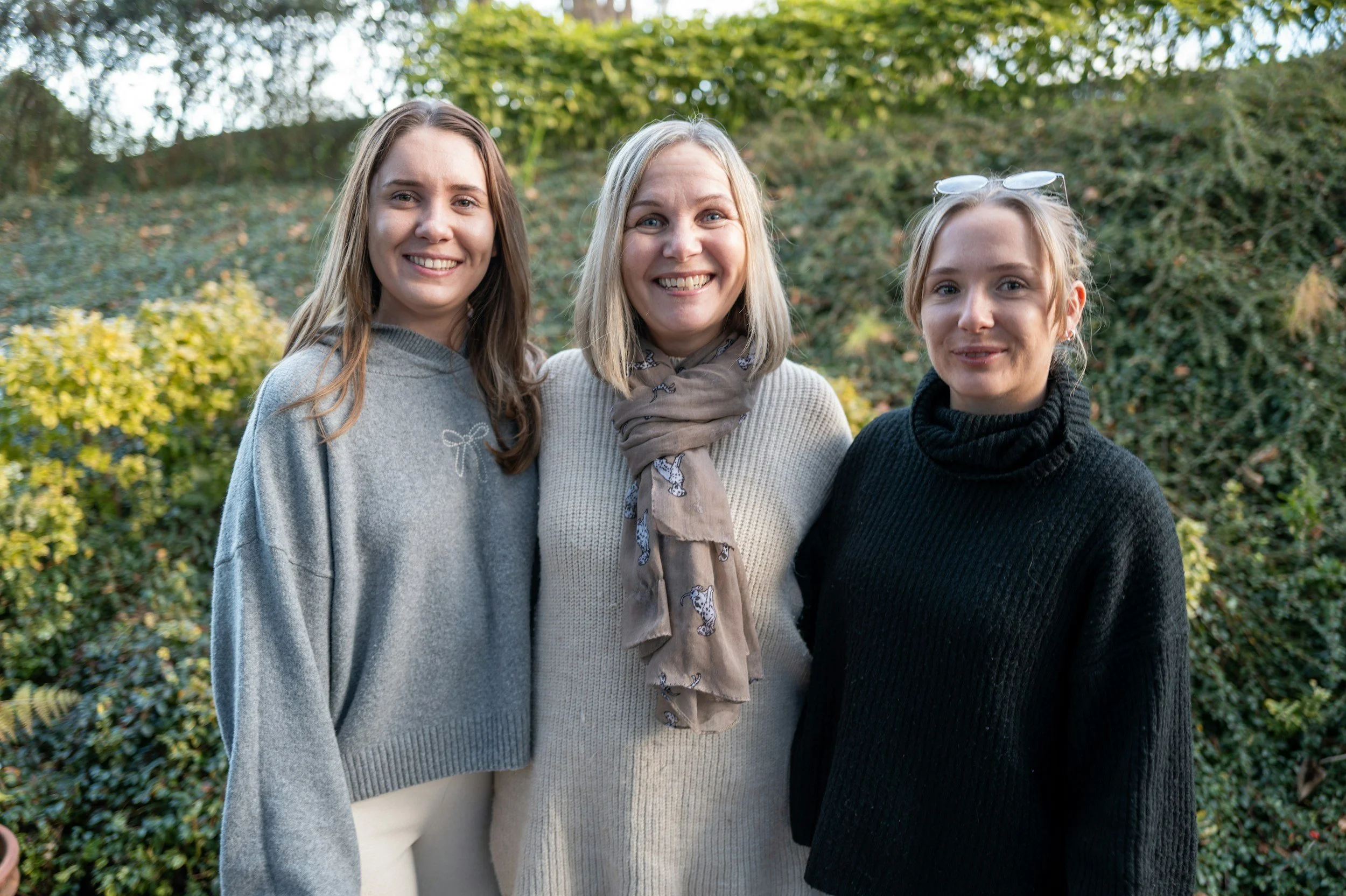 Three women standing outdoors in front of bushes, smiling at the camera. They are dressed in sweaters, with one wearing a scarf and sunglasses on her head.