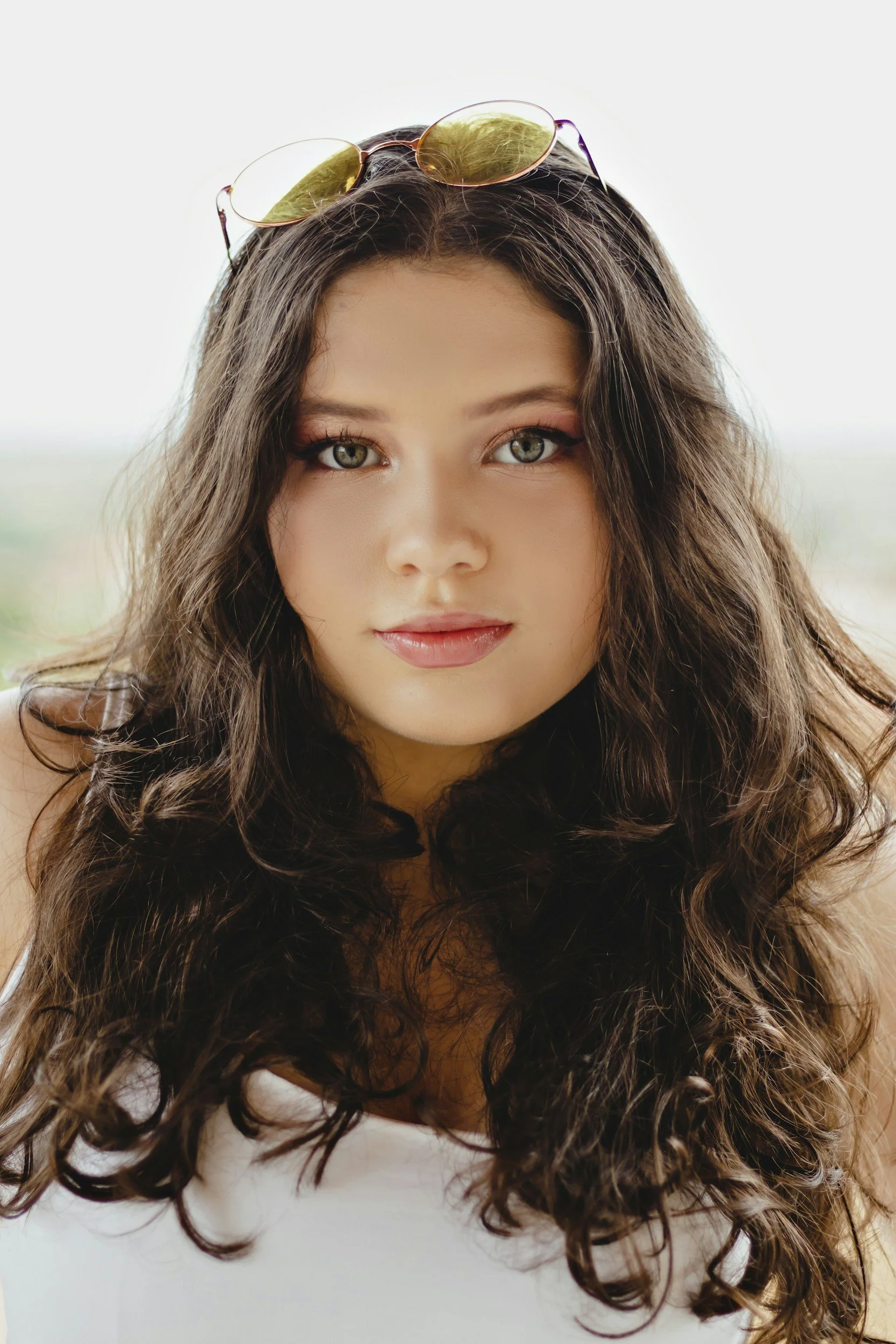 Close-up of a young woman with long, curly dark hair, light makeup, and yellow-tinted sunglasses resting on her head, looking directly at the camera.