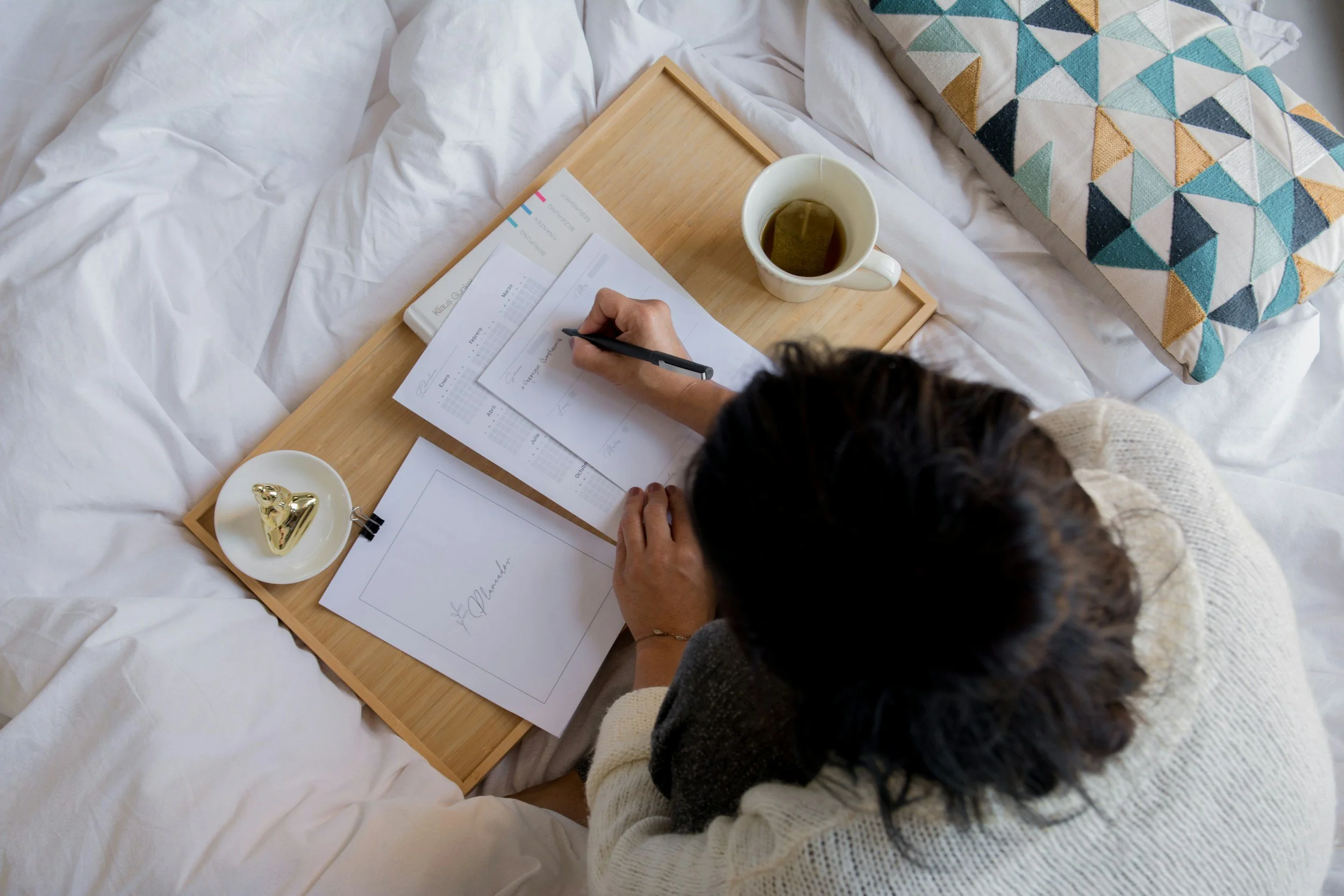 A person sitting on a bed at a wooden tray, writing on papers with a pen. There is a cup of tea, a small dish with a golden cookie, and papers on the tray. The bed has a white sheet and a patterned pillow.