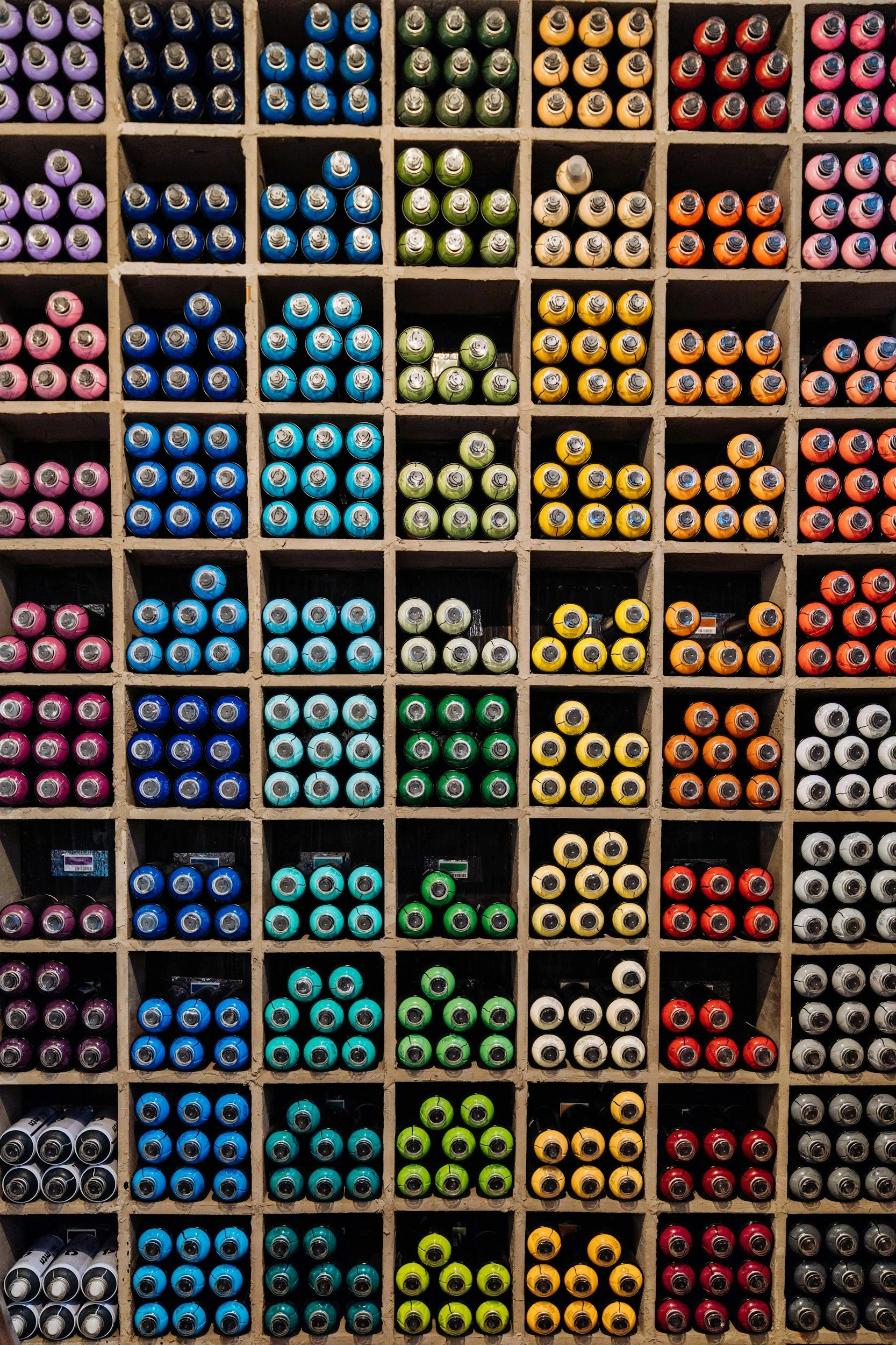 A wall of spray paint cans organized by colors, including purple, blue, green, yellow, orange, red, black, and white.