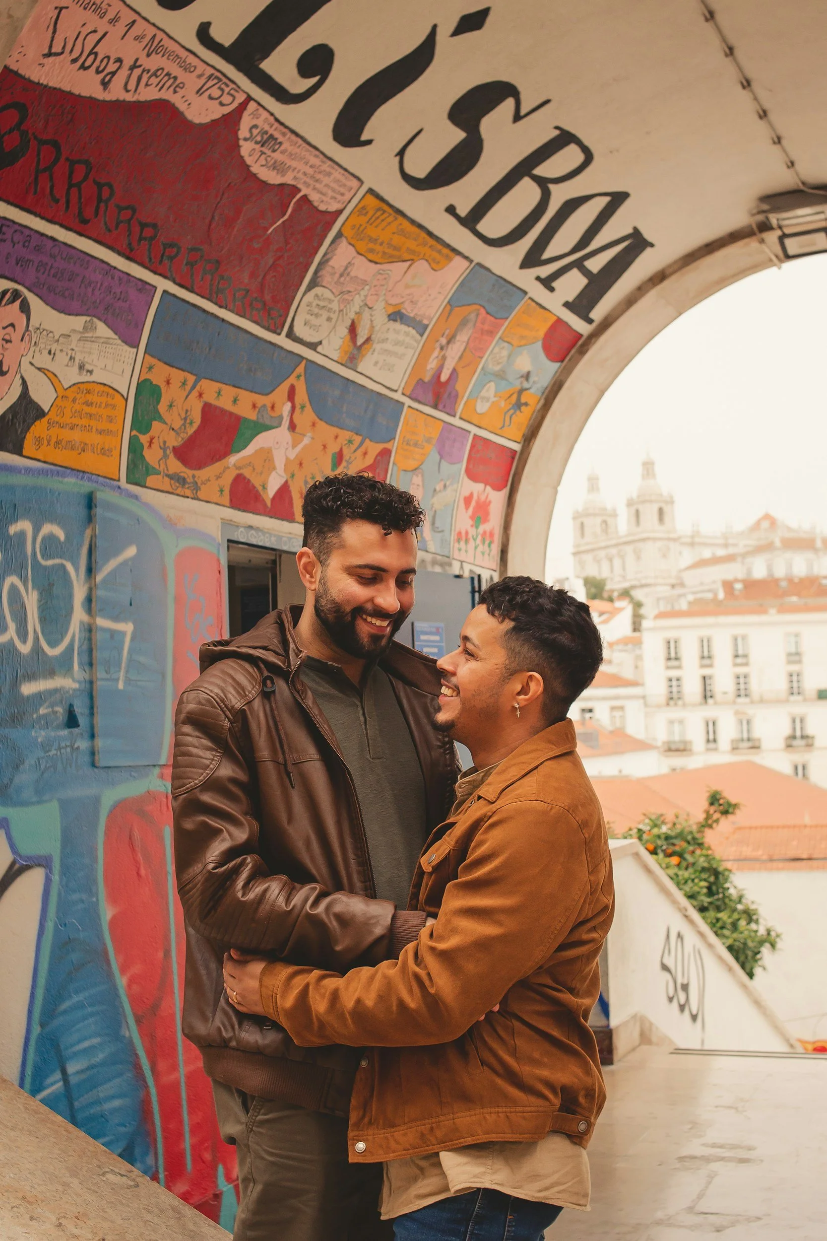 Two men smiling and looking at each other, embracing under a graffiti-covered archway with colorful murals, in an urban setting with city buildings in the background.