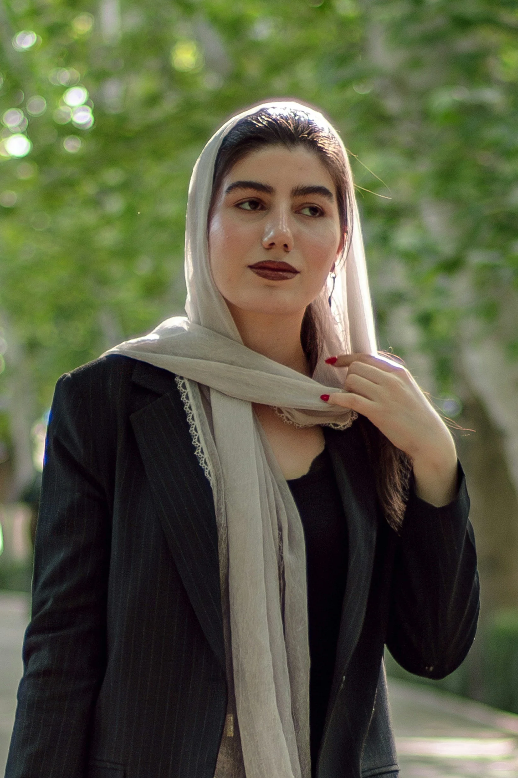 A woman with dark hair, wearing a black blazer and a beige headscarf, stands outdoors with a blurred green background, looking thoughtfully into the distance.