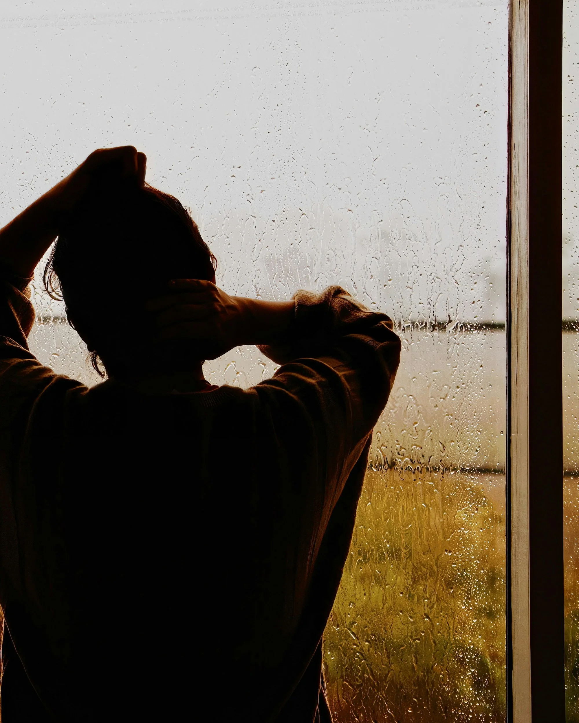 Person touching their head and neck while looking out a rain-covered window at sunset.