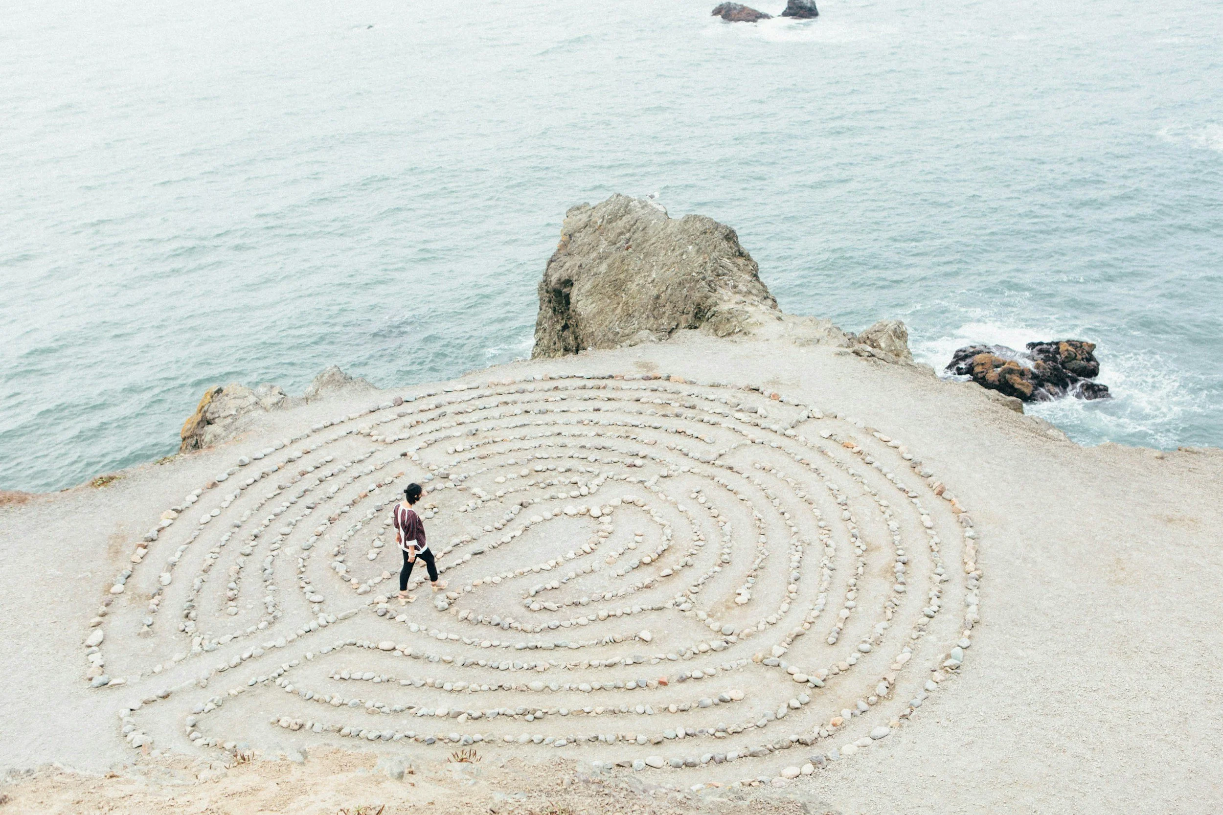A person walking through a spiral-shaped rock maze on a sandy beach by the ocean, with rocks and water in the background.