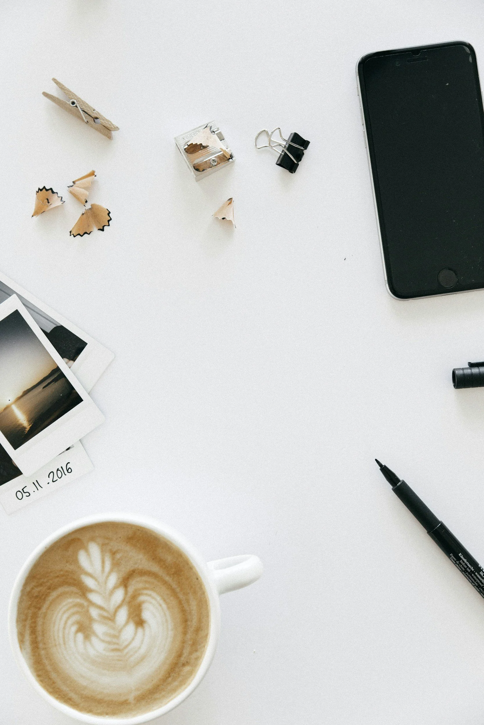 A white desk with a mug of latte, a smartphone, a black pen, a black binder clip, a small box of staples, pencil shavings, a clothespin, and Polaroid photos.