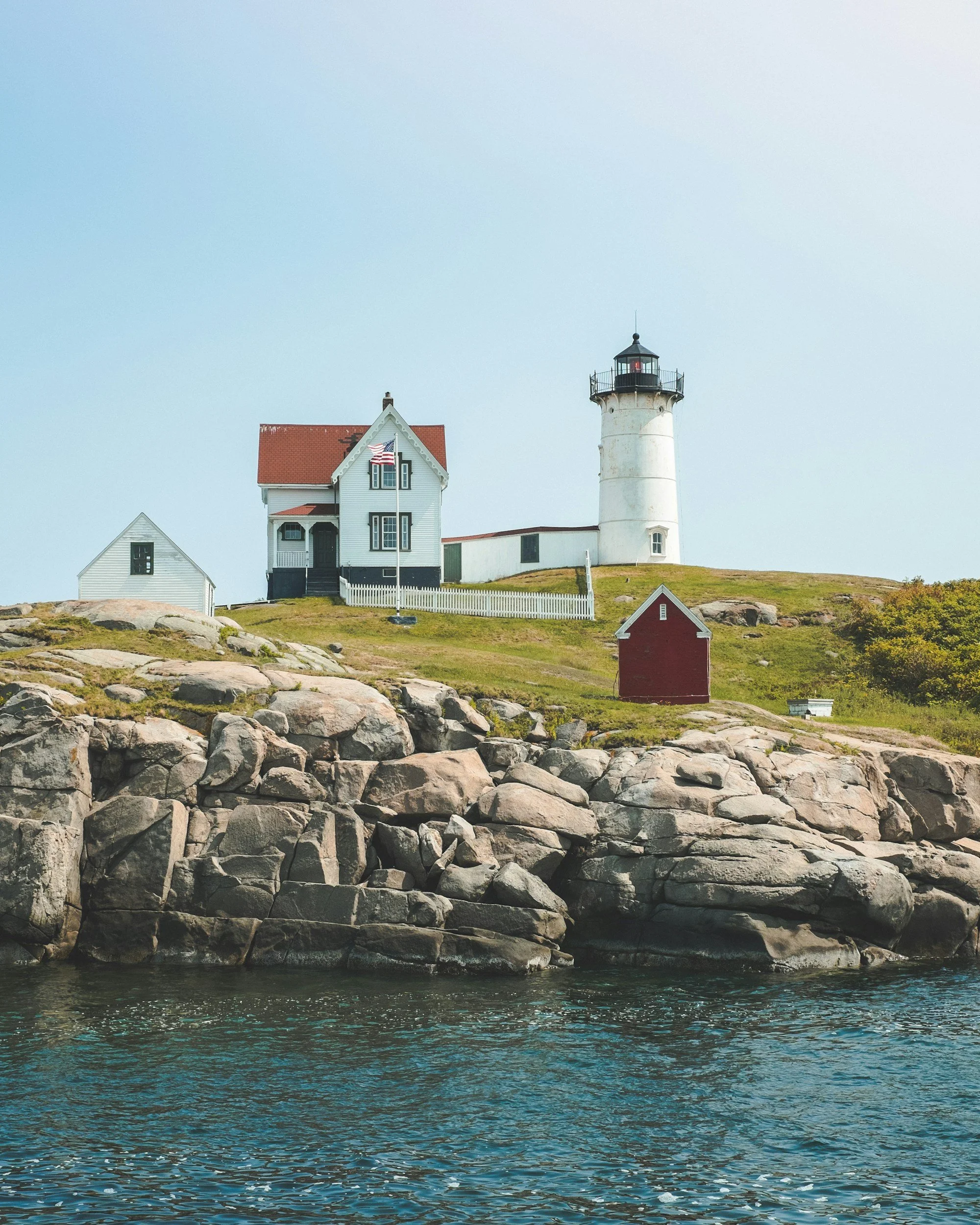 View of a lighthouse with white cylindrical structure and black lantern, a house with red roof and American flag, rocky shoreline, green grassy hill, and calm blue water under a pale sky.
