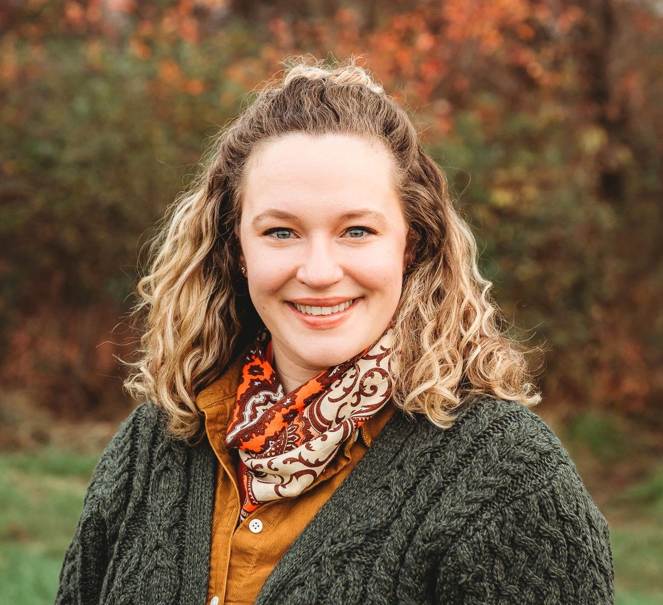 A woman with blonde curly hair smiling outdoors in fall, wearing a dark green sweater, orange shirt, and a patterned scarf.