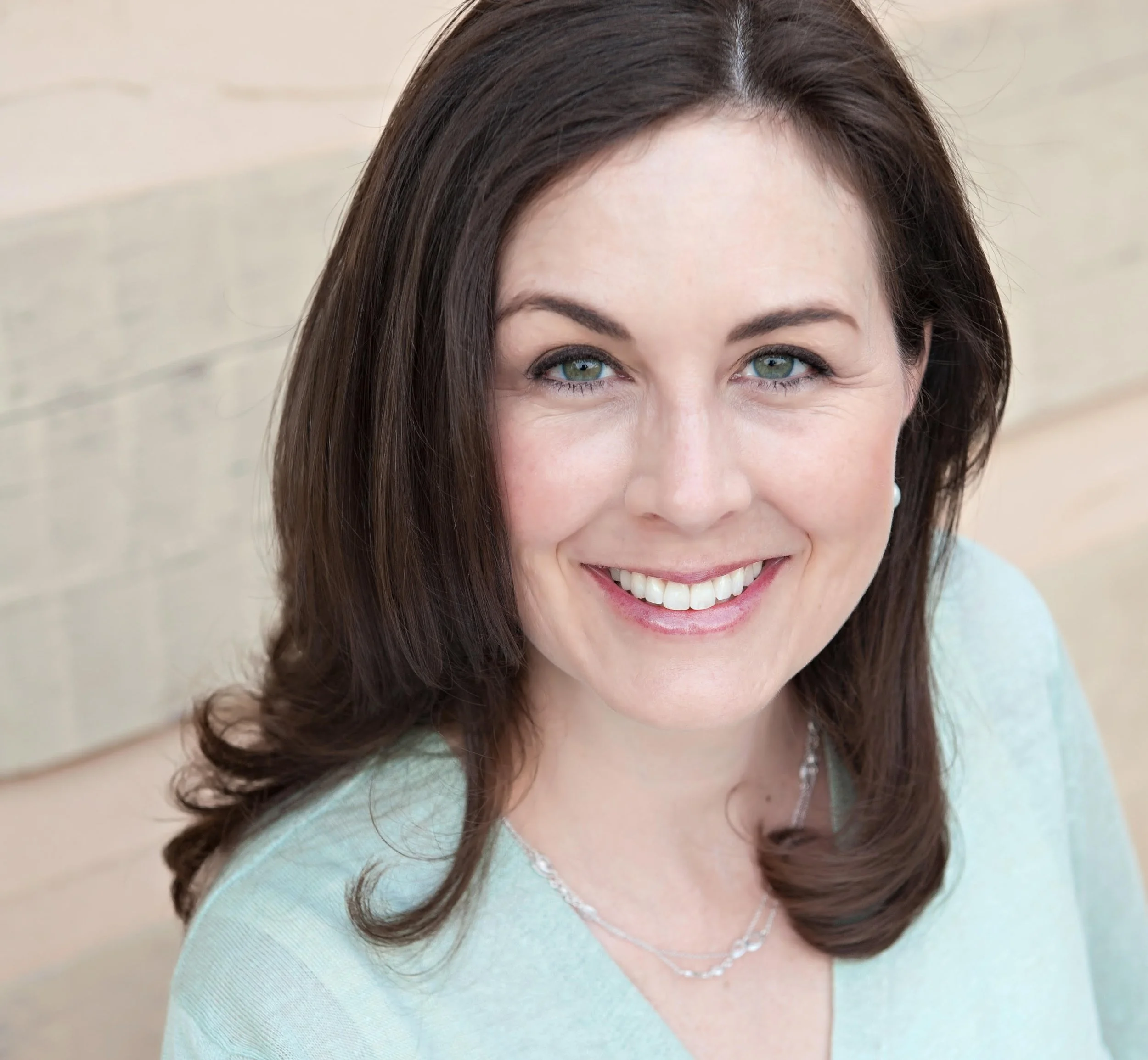Close-up of a middle-aged woman with shoulder-length brown hair, blue eyes, smiling, wearing a light blue top and pearl necklace, against a neutral background.