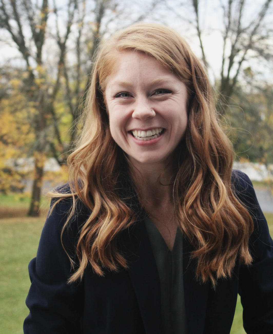 Young woman with red hair smiling outdoors in a park on an overcast day.