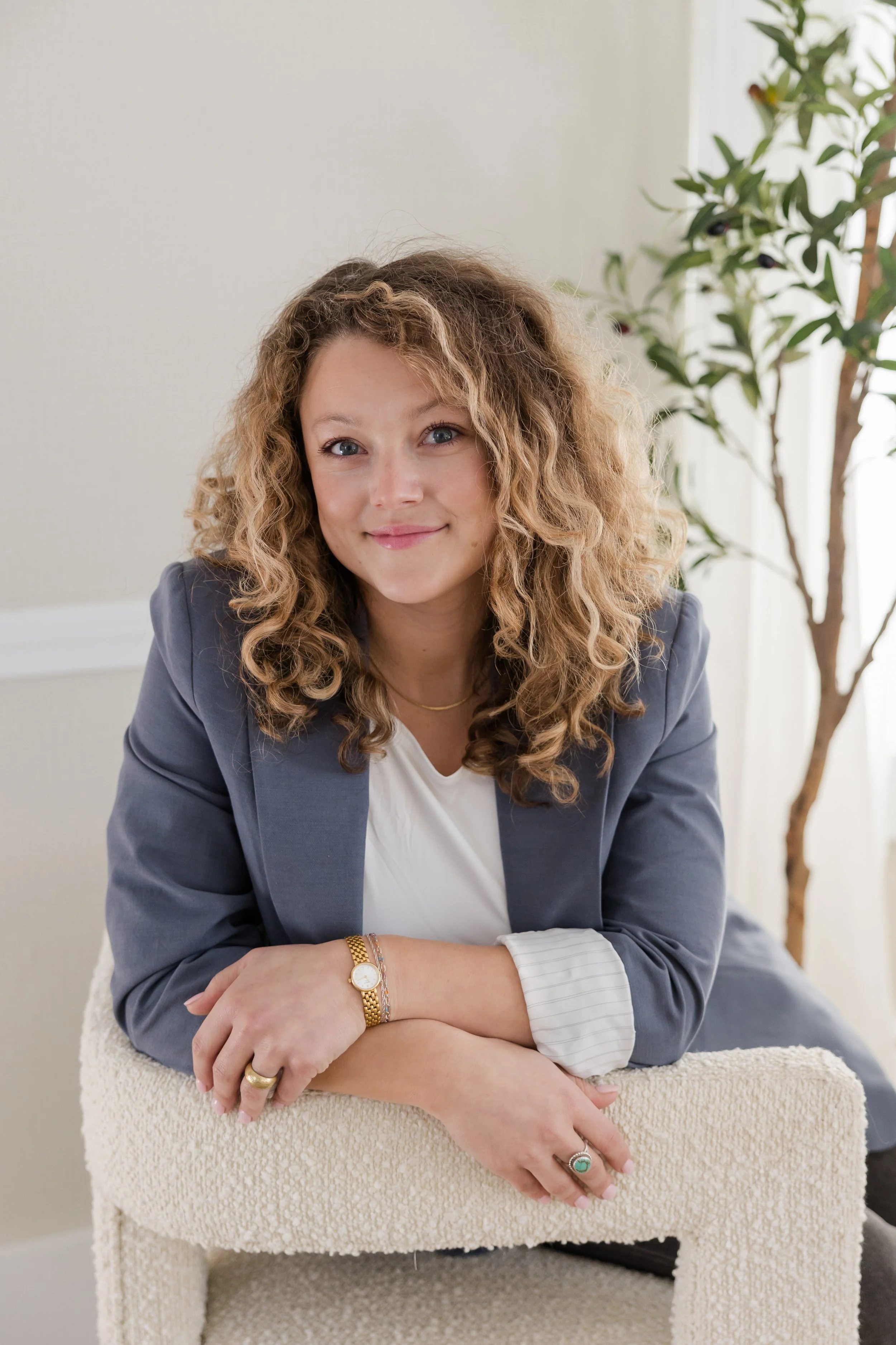 A woman with curly hair sitting on a beige textured chair in a bright room, smiling at the camera, wearing a gray blazer, white shirt, gold watch, and various rings.