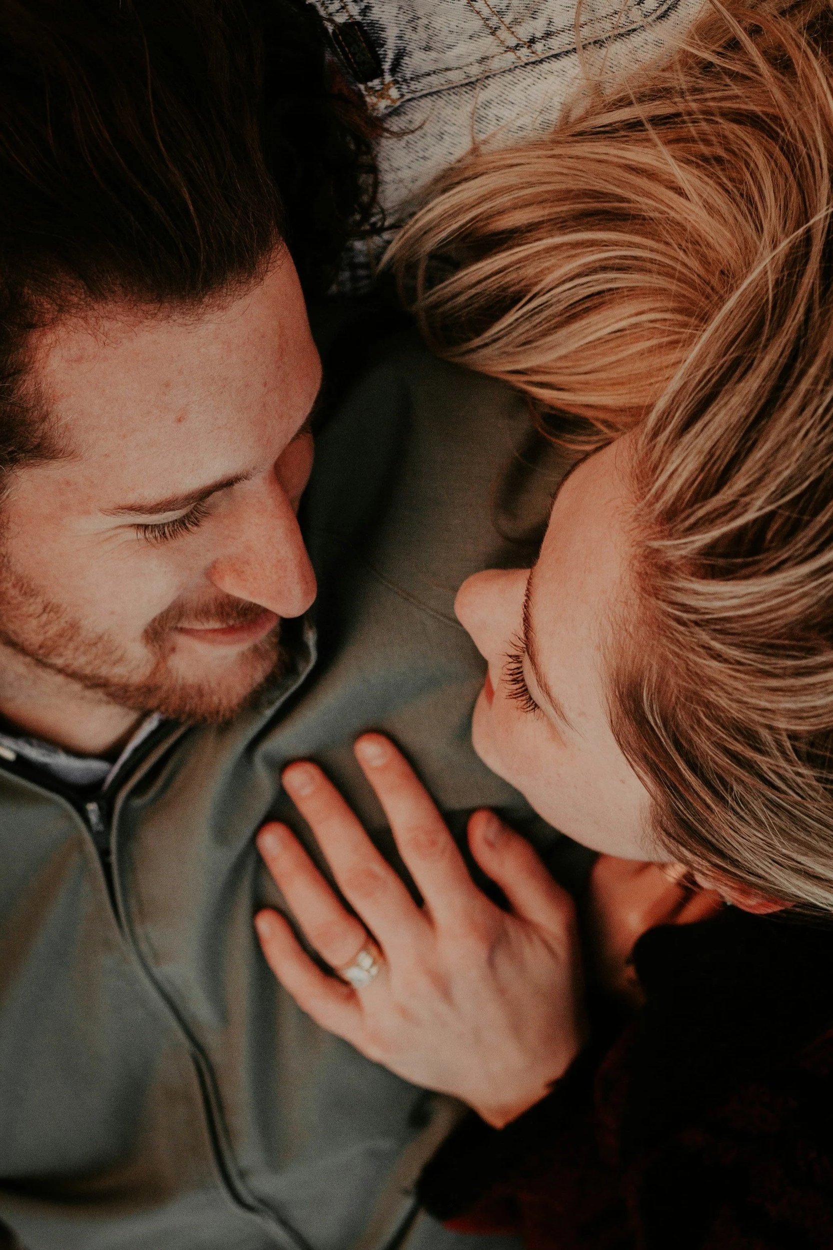 A couple lying down face to face, smiling at each other, with one woman's hand resting on the man's chest showing an engagement ring.