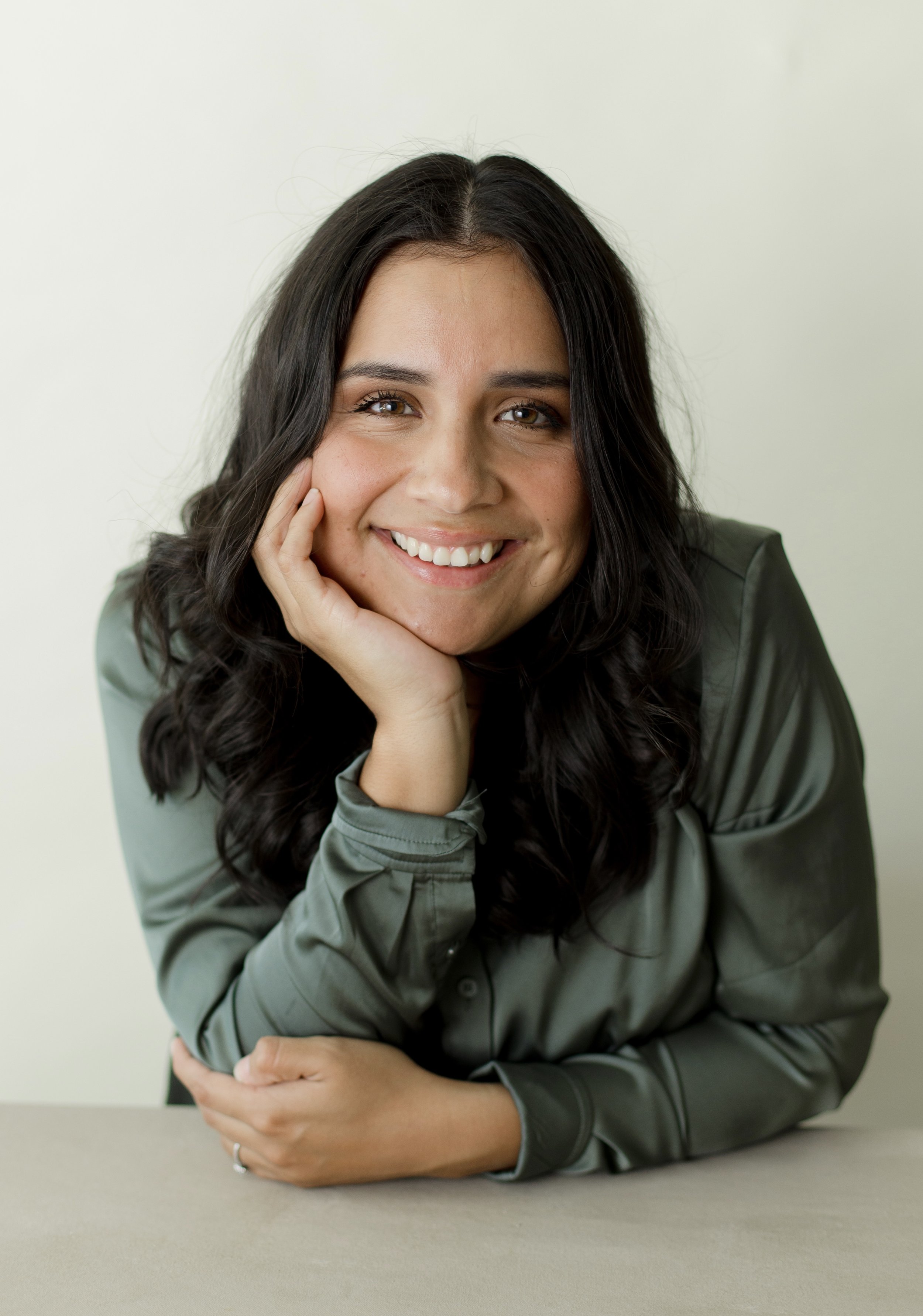 A woman with dark wavy hair smiling and resting her face on her hand, wearing a green satin shirt, sitting at a neutral-colored table against a plain light background.