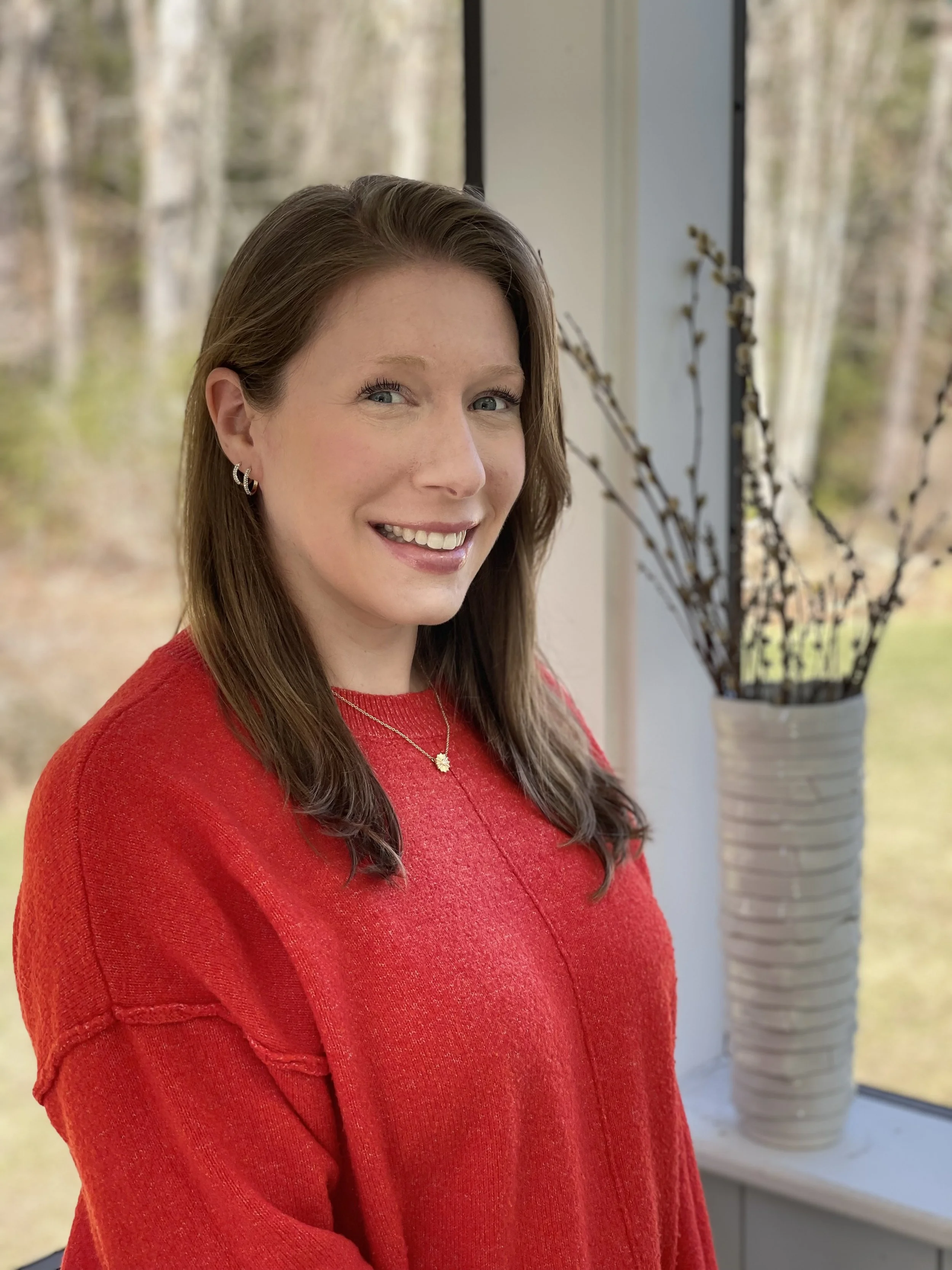 A woman with long brown hair, wearing a red sweater, standing indoors near a window with a view of trees outside.