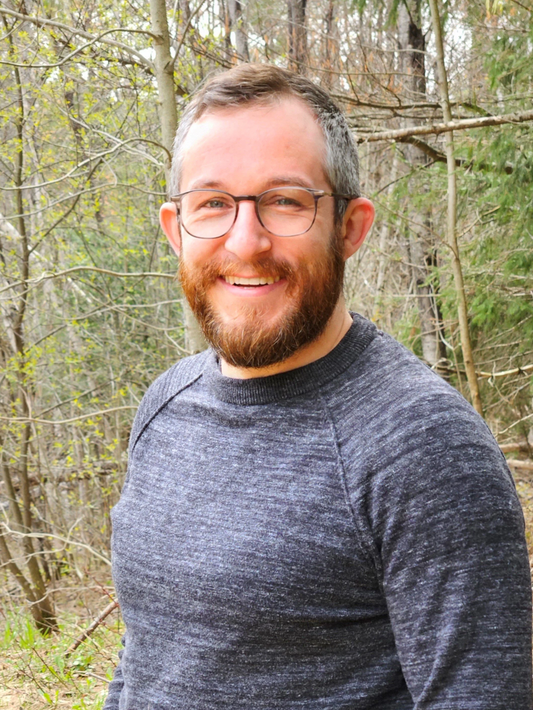 A man with glasses and a beard smiling outdoors among leafless trees and green foliage.