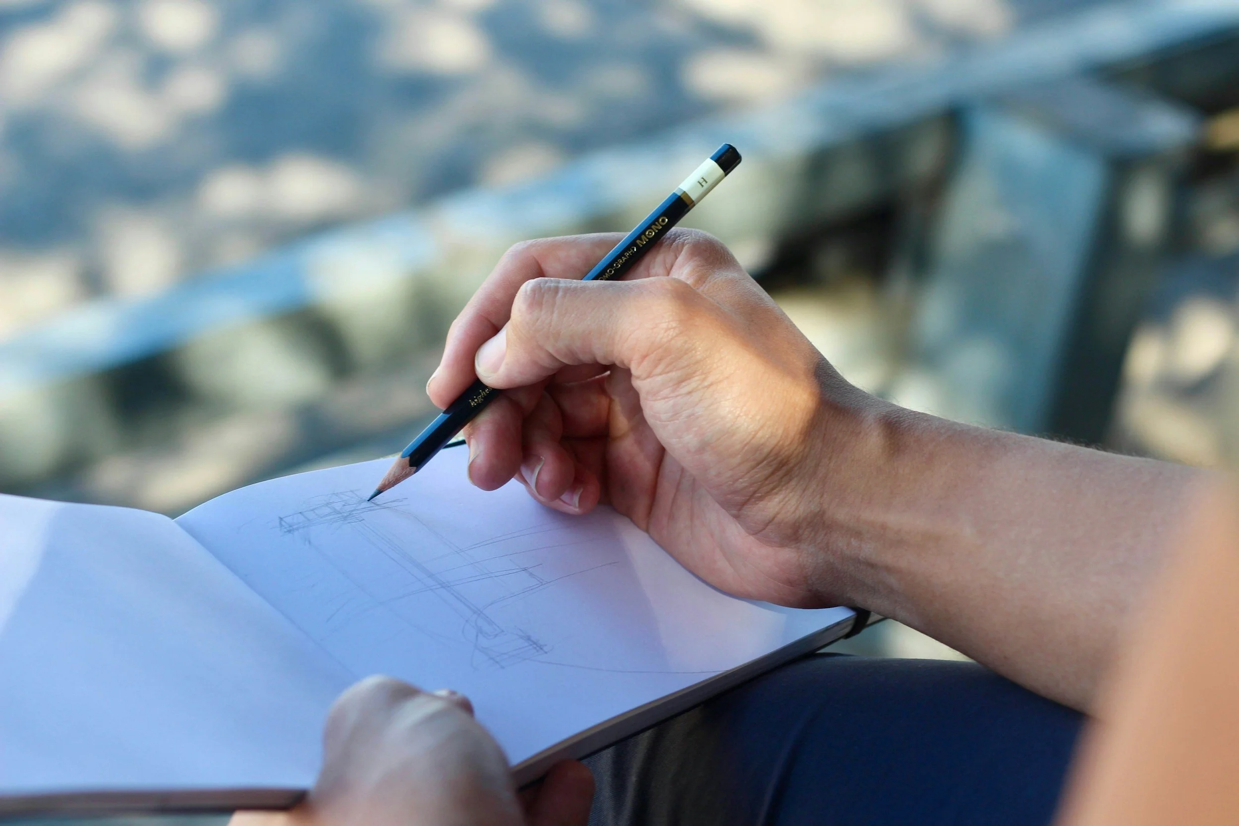 Close-up of a person's hand sketching in a notebook with a pencil, with a blurred background of a cityscape and a bridge.
