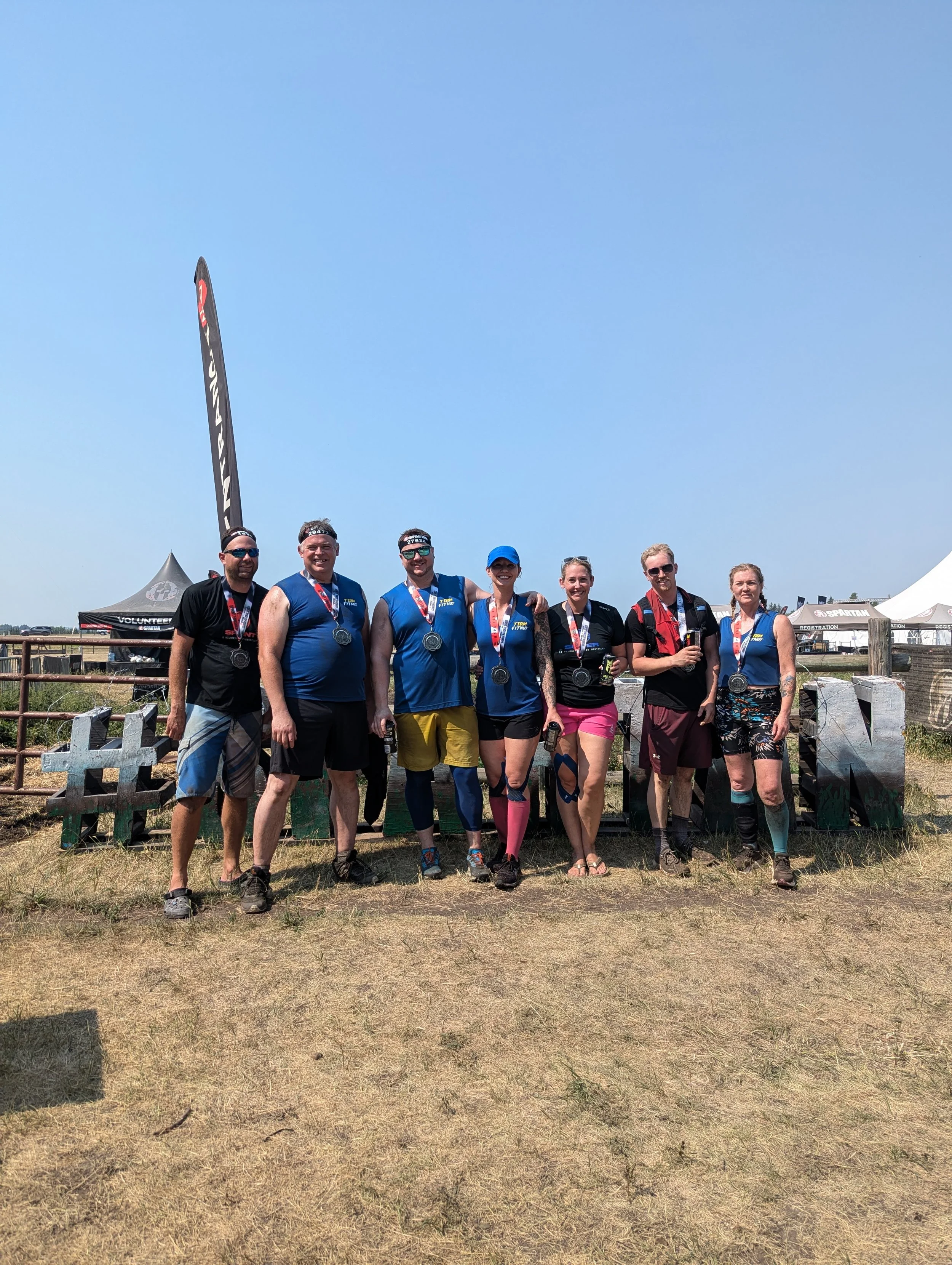 Group of seven people standing outdoors with medals around their necks, posing for a photo at an outdoor event.