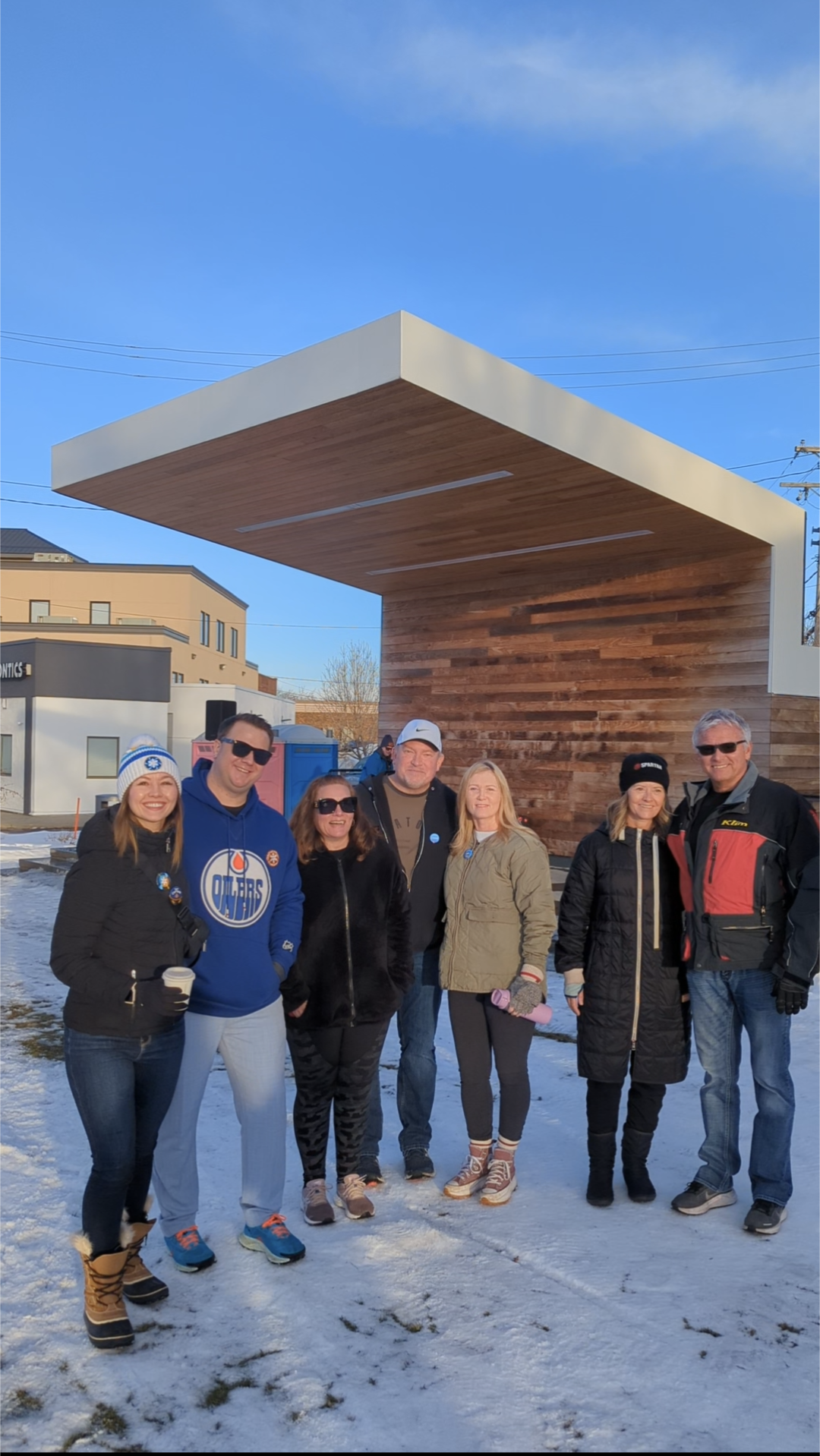 Group of seven people standing outdoors in winter, dressed warmly, in front of a modern wooden structure with a large cantilevered roof, with snow on the ground and a clear blue sky.