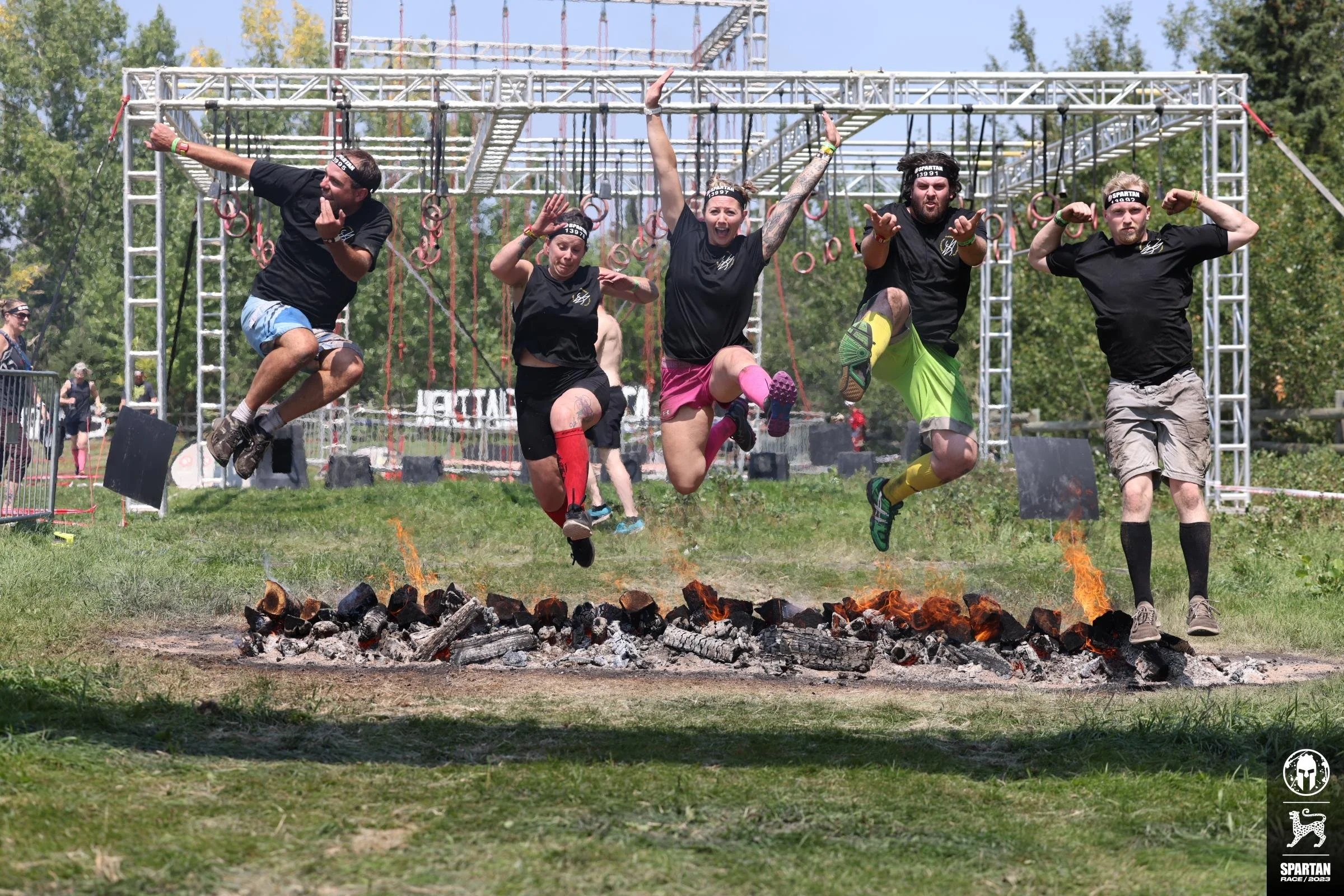 Group of five people jumping over fire on a Spartan Race obstacle course during daytime, with metal scaffolding and trees in the background.