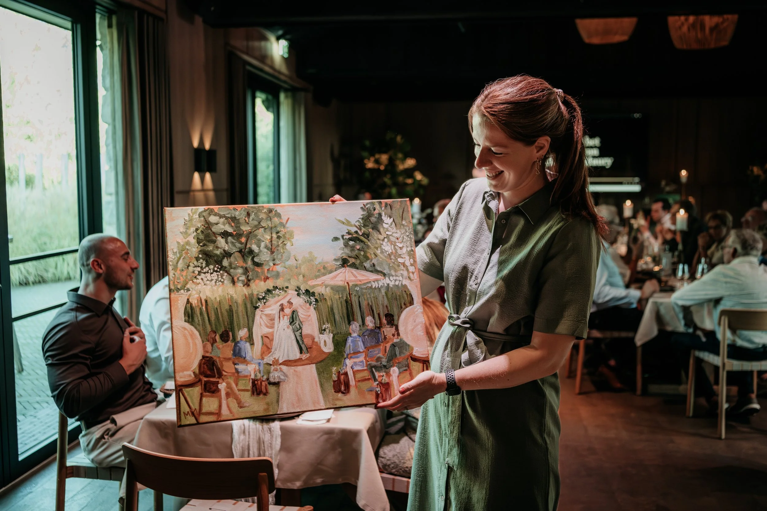 Vrouw toont een schilderij aan een groep mensen tijdens een bijeenkomst in een restaurant of zaal.
